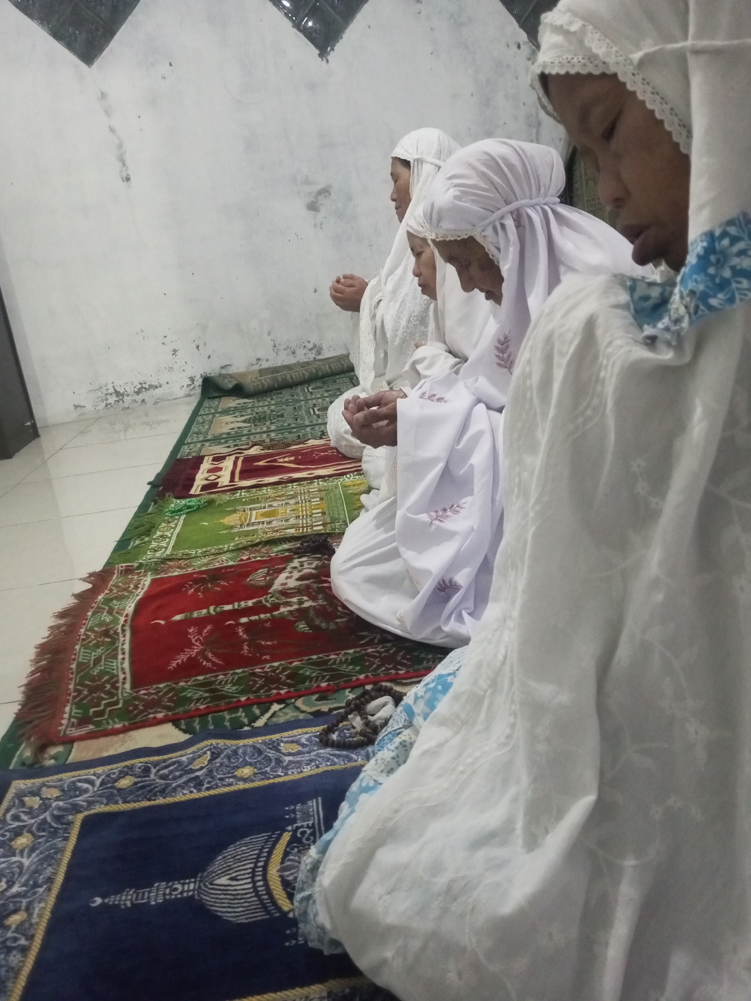 In a dimly lit room, four individuals, likely female, are seated on prayer mats facing the same direction, engaged in prayer. They are all wearing white hijabs and modest white clothing. The prayer mats are arranged in a row, with colorful geometric patterns. The walls of the room appear to be white and slightly textured, with some imperfections. A tiled floor is visible in the foreground, and a faint shadow of what might be a window or opening can be seen on the wall. The overall atmosphere suggests a religious observance. The image was taken in Andongsili, Indonesia.
