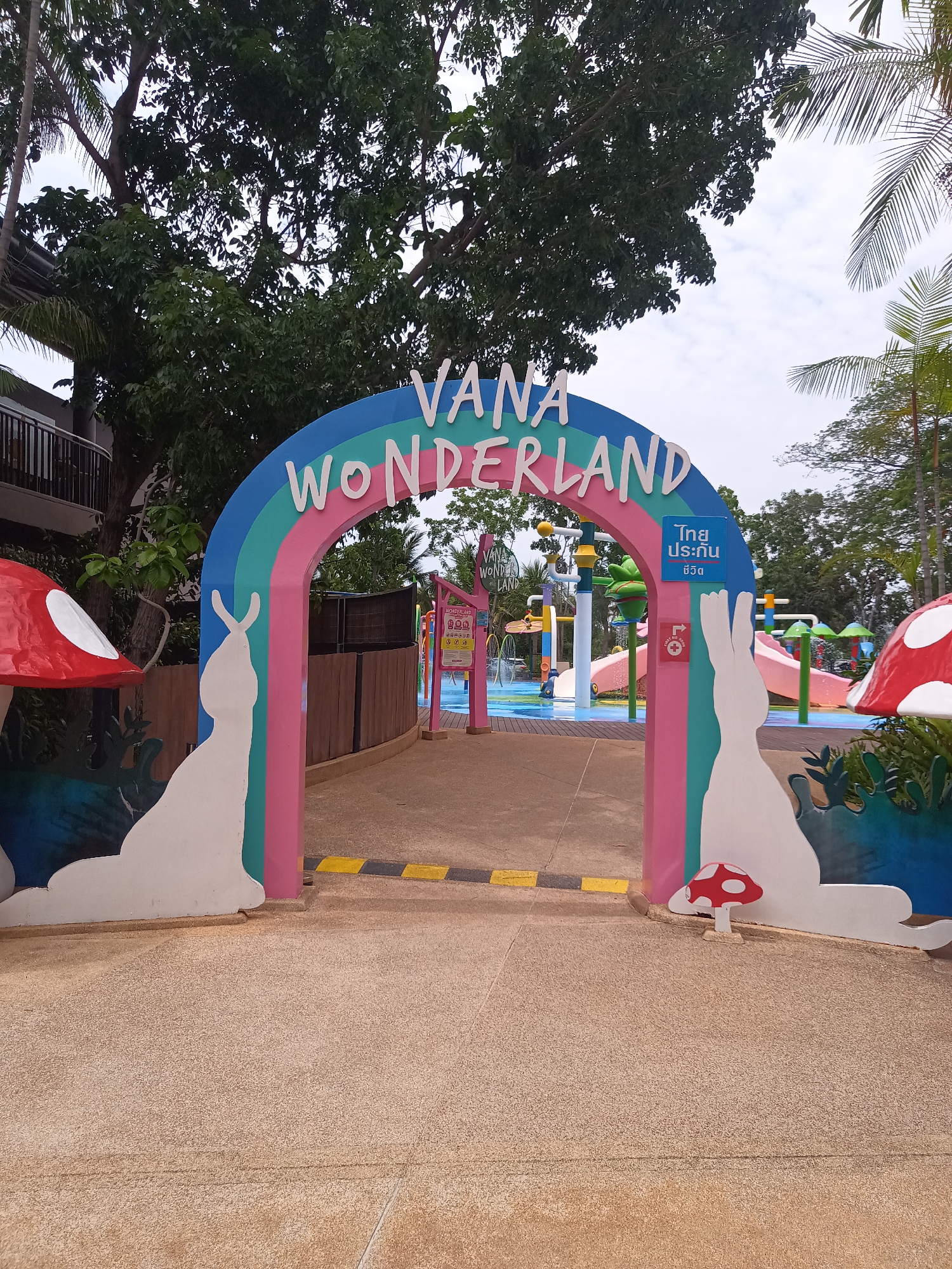 The image shows the entrance to Vana Wonderland in Hua Hin City Municipality, Thailand. The entrance is marked by a large, colorful arch with the words "Vana Wonderland" written on it. The arch is decorated with rabbit silhouettes and mushroom figures. In the background, there is a water park with slides and other attractions. There are no people visible in the image. The sky is overcast, and the surrounding environment includes trees and buildings.