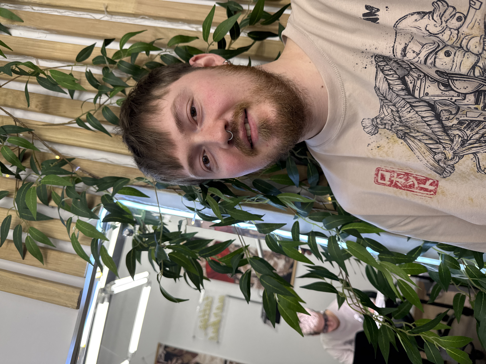 A Caucasian male with a beard and a nose ring is shown from the chest up. He is wearing a beige t-shirt with a graphic design on it. The background consists of a wooden-slatted wall with green leafy vines intertwined. Part of a mirror and some lighting fixtures are visible. The location appears to be inside a commercial establishment, possibly a salon or studio. The location is Chaikovsky, Russia.