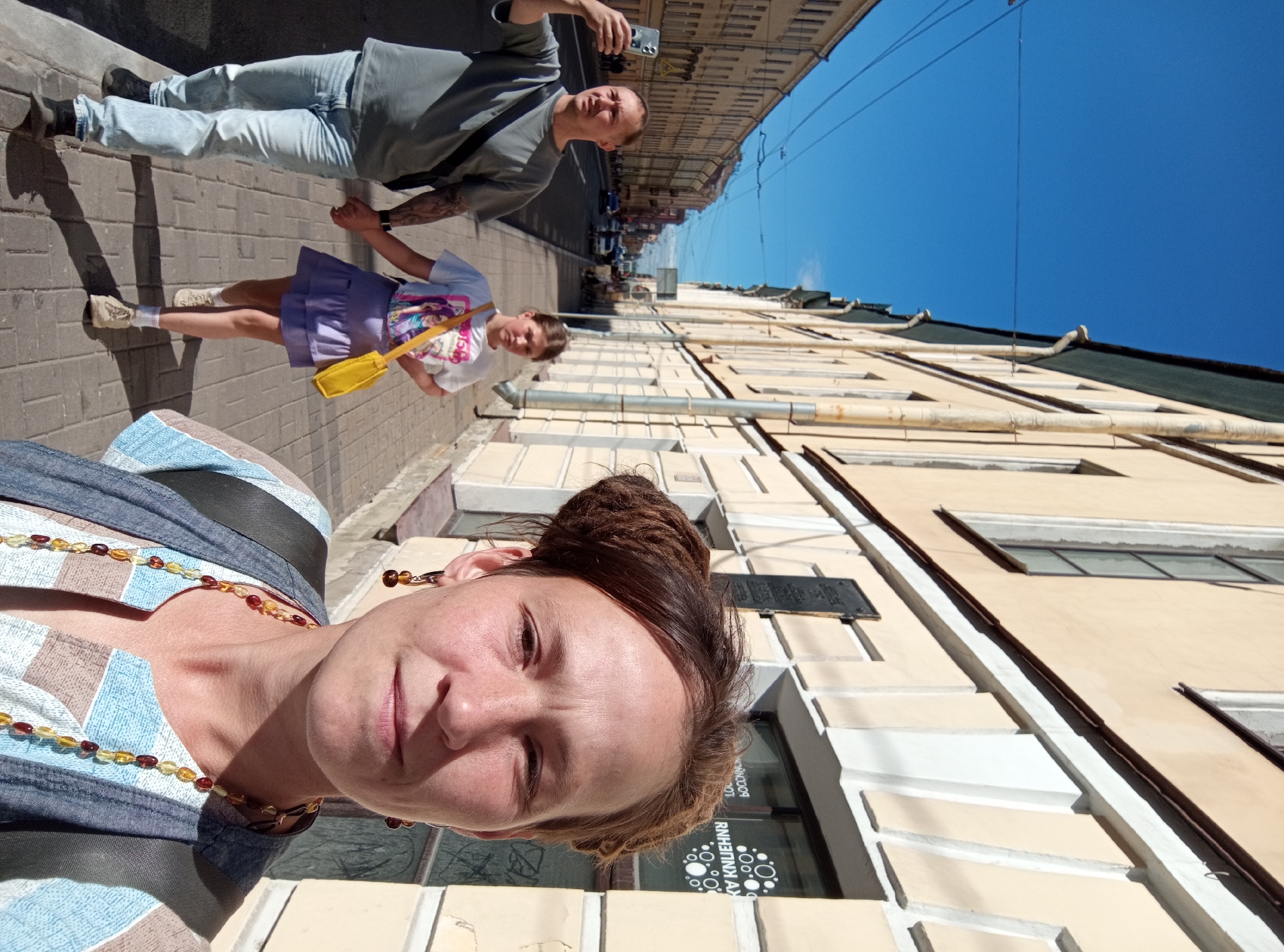 This is a selfie taken by a woman in St. Petersburg, Russia, on a sunny day. She is in the foreground, with her face close to the camera and her hair pulled up in a bun. She is wearing a blue and white striped top and a beaded necklace. Behind her, a man and a young girl are walking away from the camera. The man is wearing a grey t-shirt and jeans, and the girl is wearing a purple skirt and a white t-shirt. They are walking on a sidewalk lined with buildings. The buildings appear to be old and ornate, with cream-colored facades and dark roofs. There are trees and streetlights visible along the sidewalk. The sky is a clear blue. The overall mood of the image is casual and relaxed.