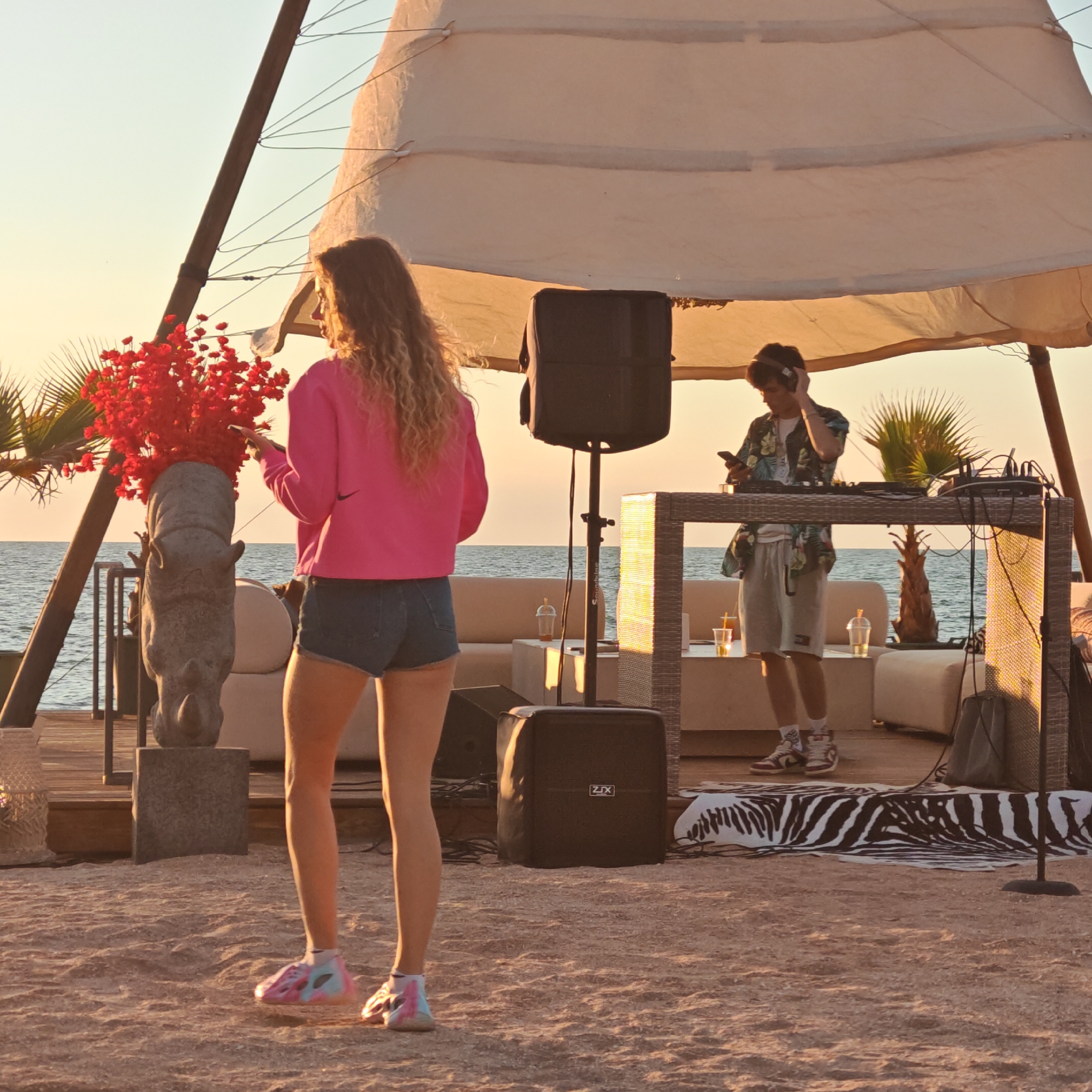 The image depicts an outdoor scene with a DJ set up on a raised platform, playing music. A person with headphones is standing behind a DJ console, holding a phone. In the foreground, a woman with long, curly blonde hair is facing away from the camera, wearing a pink sweatshirt and denim shorts. She is holding a bouquet of red flowers. The setting appears to be a beach or coastal area, with the sea visible in the background. There are lounge chairs and other decorative elements suggesting a party or event. The lighting indicates it is either sunrise or sunset. The location is specified as Gorodskoye poseleniye Shchelkino, Ukraine, which is an urban-type settlement in Crimea.