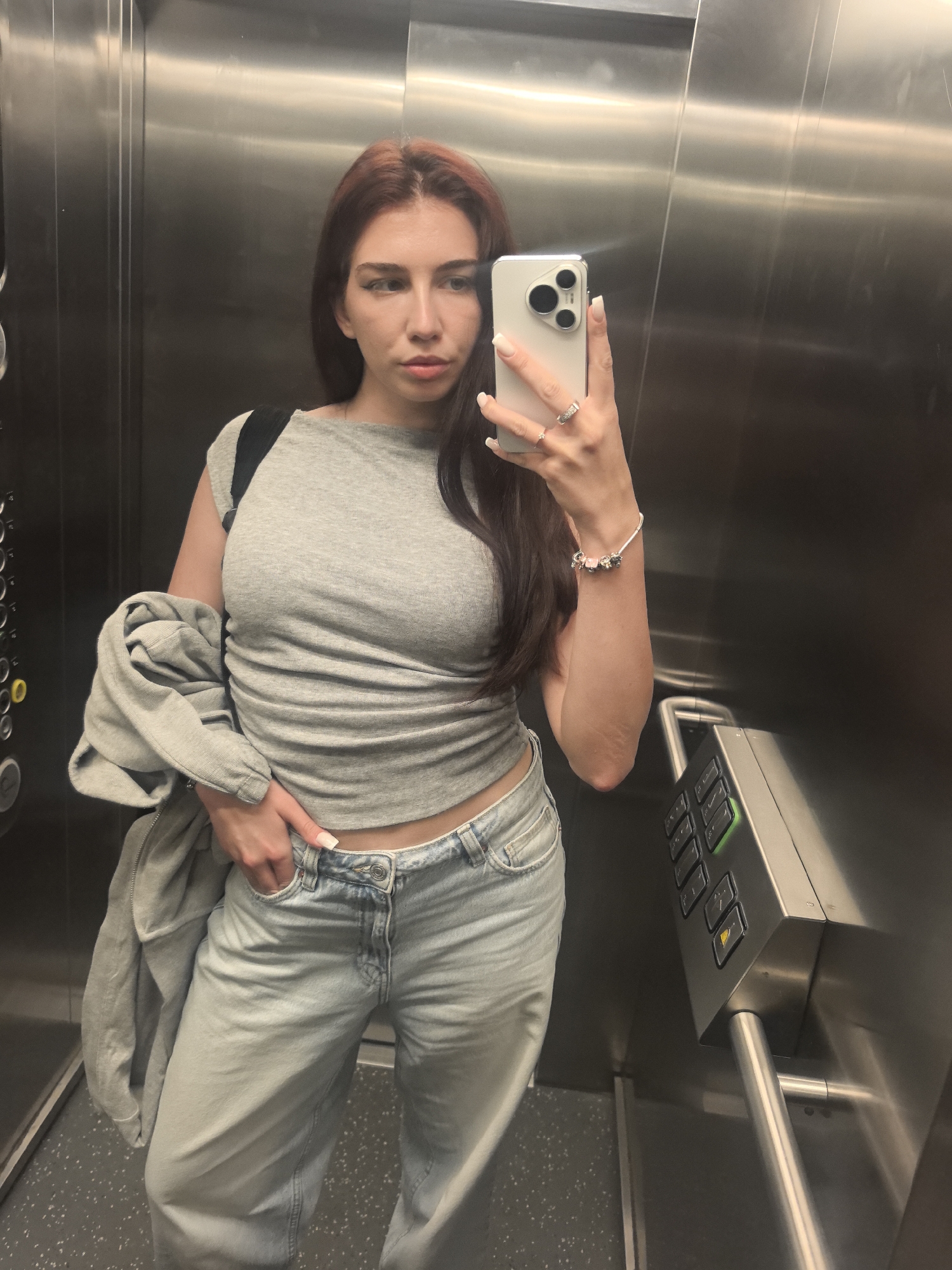 A young woman is shown taking a selfie in a reflective elevator interior. She is positioned centrally, gazing directly into the camera of the light-colored smartphone held in her right hand. Her long, dark reddish-brown hair is parted in the middle and falls over her shoulders. Her expression is neutral, with no discernible smile.
She is dressed in a light grey, short-sleeved crop top that reveals a sliver of her midriff, and light-wash denim jeans. A grey jacket or hoodie is casually draped over her left arm, and a black backpack strap is visible on her left shoulder. Her left hand is tucked into the pocket of her jeans. She wears multiple rings on her right hand and a charm bracelet on her left wrist, and her fingernails have white tips.
The elevator's interior features highly polished, metallic walls, likely stainless steel, which reflect her image. The floor is dark with a speckled pattern. On the left wall, a panel with numerous elevator floor buttons is visible, some illuminated in yellow (for 'C') and green. Another control panel is on the right, also with illuminated green buttons, directional arrows, and door controls, situated above a horizontal metal grab bar. The image captures an everyday moment within a building, specifically an elevator, in Bogomilovo, Bulgaria. The time of day and weather conditions are not evident.