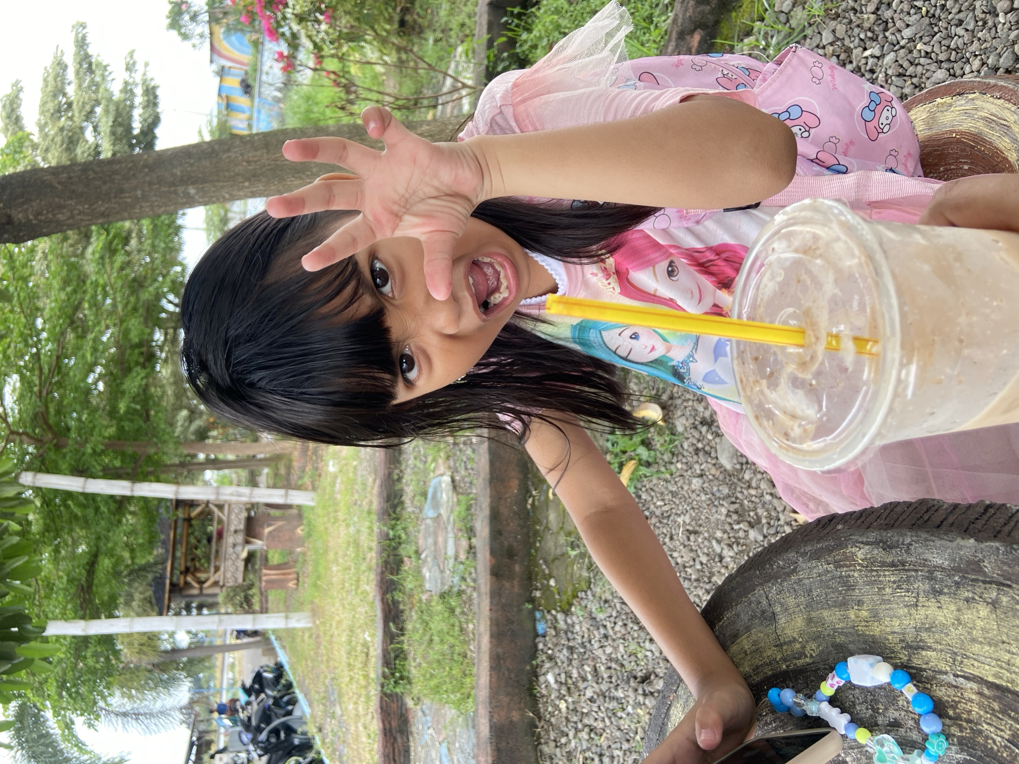 A young girl is prominently featured, playfully extending one hand towards the camera with fingers spread, while holding a large plastic cup containing a brown beverage with a yellow straw in the other hand. She's wearing a light pink dress-like top with cartoon characters. The child appears to be sitting, perhaps on a decorative structure that looks like the end of a log or tree.
In the background, there's a slightly blurred outdoor environment. There is some green grass, foliage, and trees. There is also what appears to be a gazebo structure, a line of parked motorcycles, and a multi-colored wall feature.
This scene is located in Kalitengah, Indonesia.