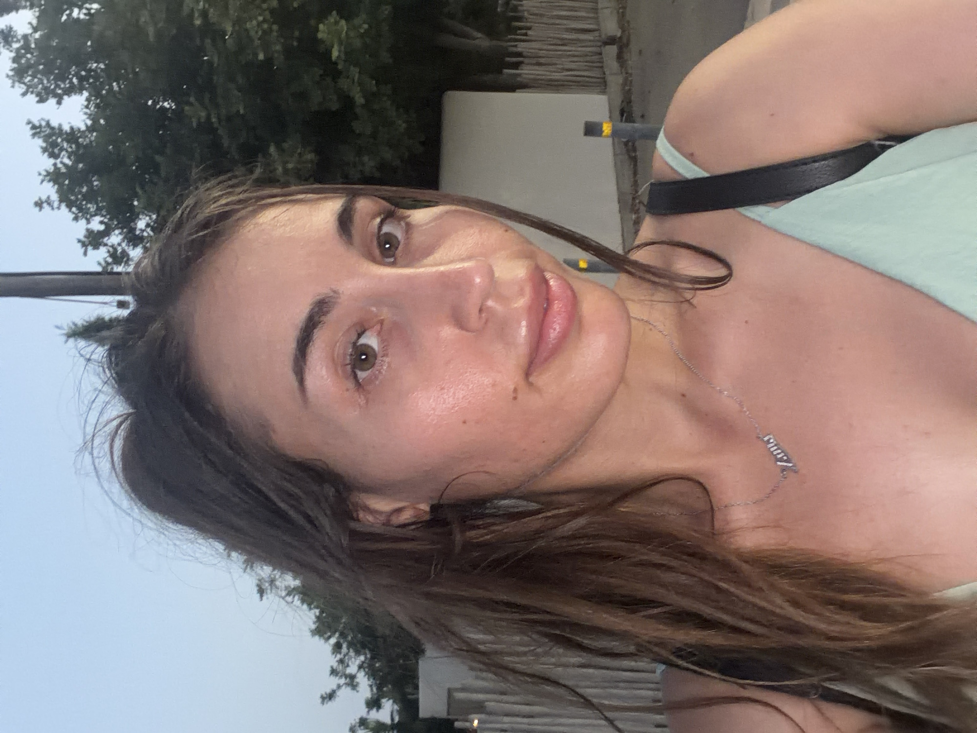 A young woman with long, dark brown hair and brown eyes is pictured in a close-up, outdoor selfie. She is wearing a light mint green strap top and a silver necklace with a nameplate. Her skin appears slightly shiny, possibly from sweat or a dewy makeup product. She is outdoors, with trees and a building visible in the background. The lighting suggests it is either dawn or dusk. The location is identified as Pervolia, Cyprus, indicated by a visible street sign with the name "Pervolia."