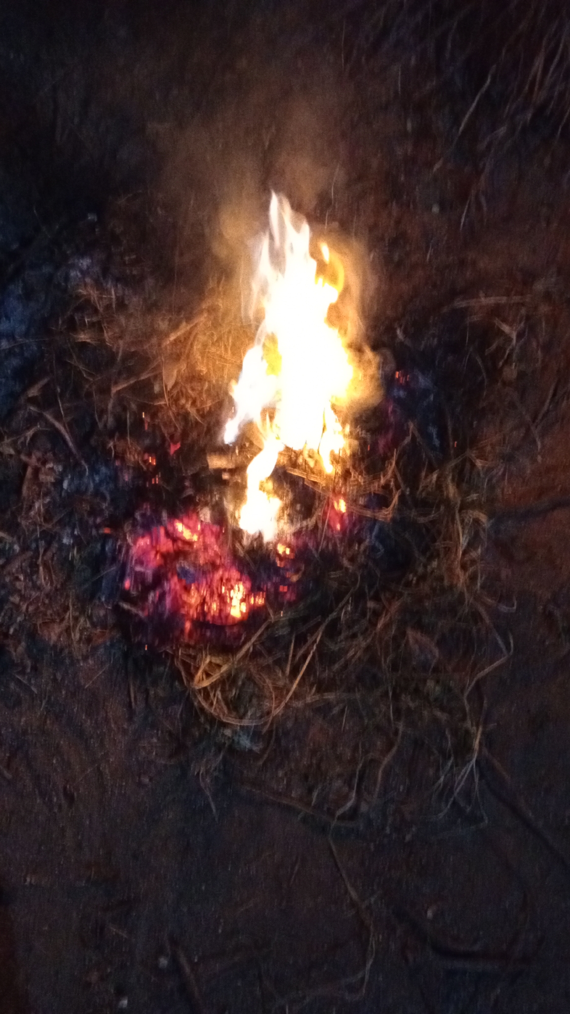The image depicts a small, open fire burning in what appears to be an outdoor area. The fire is centrally located and consists of bright yellow and white flames at its core, transitioning to red and orange hues at the base, suggesting embers and burning material. Surrounding the fire is a pile of dry, straw-like debris, along with some darker, possibly charred material. Wisps of smoke are visible rising from the flames and dispersing into the darker background. The ground around the fire is dark and appears to be earth or dirt, with some scattered dry vegetation. The overall lighting suggests it is either nighttime or dusk, with the fire being the primary light source. There are no people visible in the image. The image's context specifies it is in Buketteukuh, Indonesia, which suggests an outdoor setting, possibly a rural or semi-rural location.