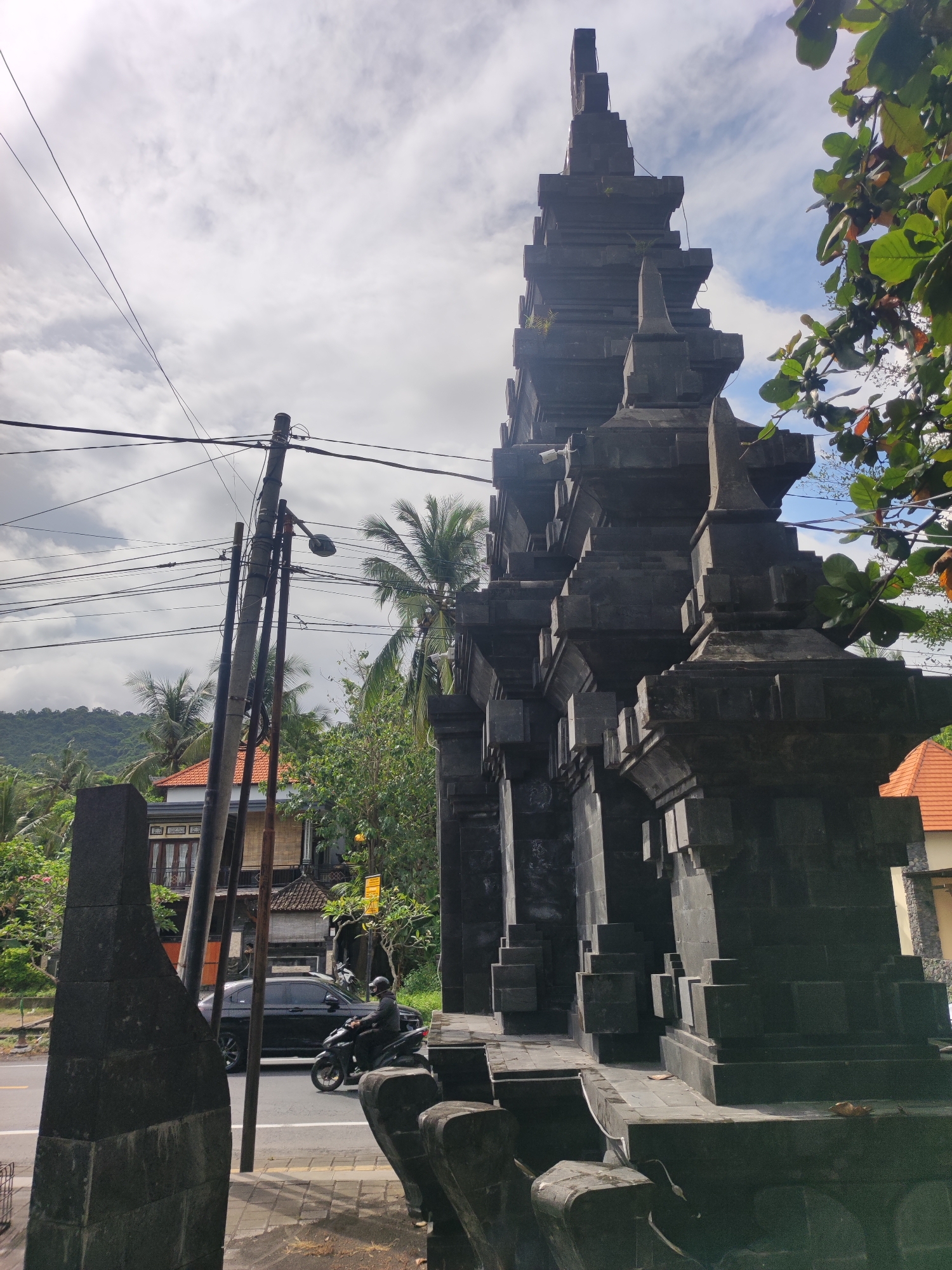 A ground-level, daytime shot captures a weathered, gray stone Balinese gate in Padangbai, Indonesia.
A motorcyclist wearing a helmet rides past the gate on a paved road with a black car driving behind. The gate has a multi-tiered, stepped structure. Power lines are visible, stretching over the road and an adjacent utility pole. A building with a red-tiled roof is on the right side of the gate. In the background, dense tropical vegetation with palm trees covers a hill. The sky is cloudy with patches of blue.