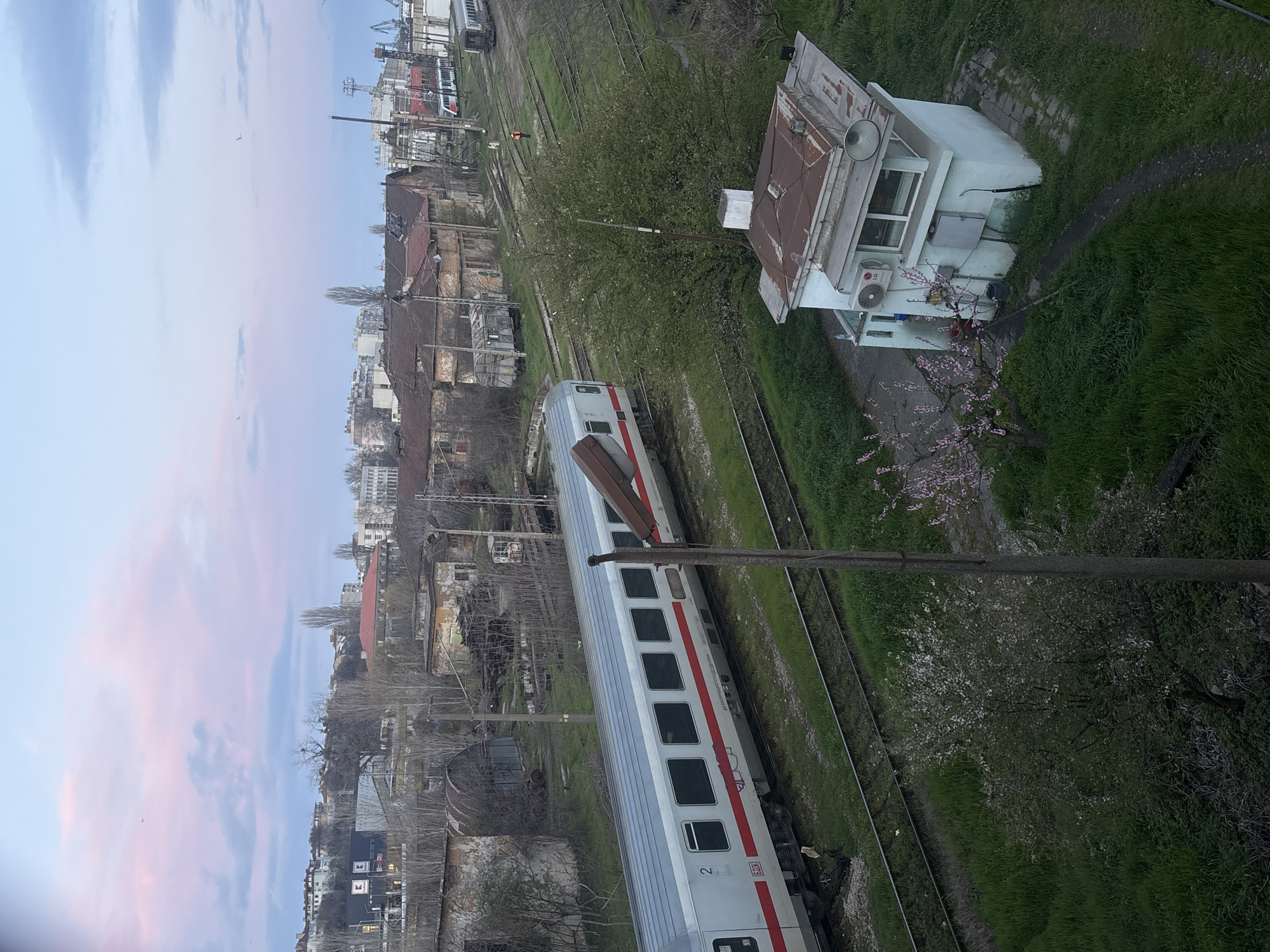 The image shows a high-angle view of a railway scene in Varna, Bulgaria. A passenger train, with a silver exterior and red accents, is stopped on the tracks. Adjacent to the tracks is a small, weathered control building with a rusty roof, an external speaker, and an LG air conditioning unit. A cherry blossom tree is in full bloom nearby. Various other tracks are visible, some leading towards industrial port facilities in the background. Derelict buildings and overgrown vegetation are scattered around the area. In the distance, the cityscape of Varna fades into the horizon under a sky tinged with pink hues. A utility pole extends into the foreground.