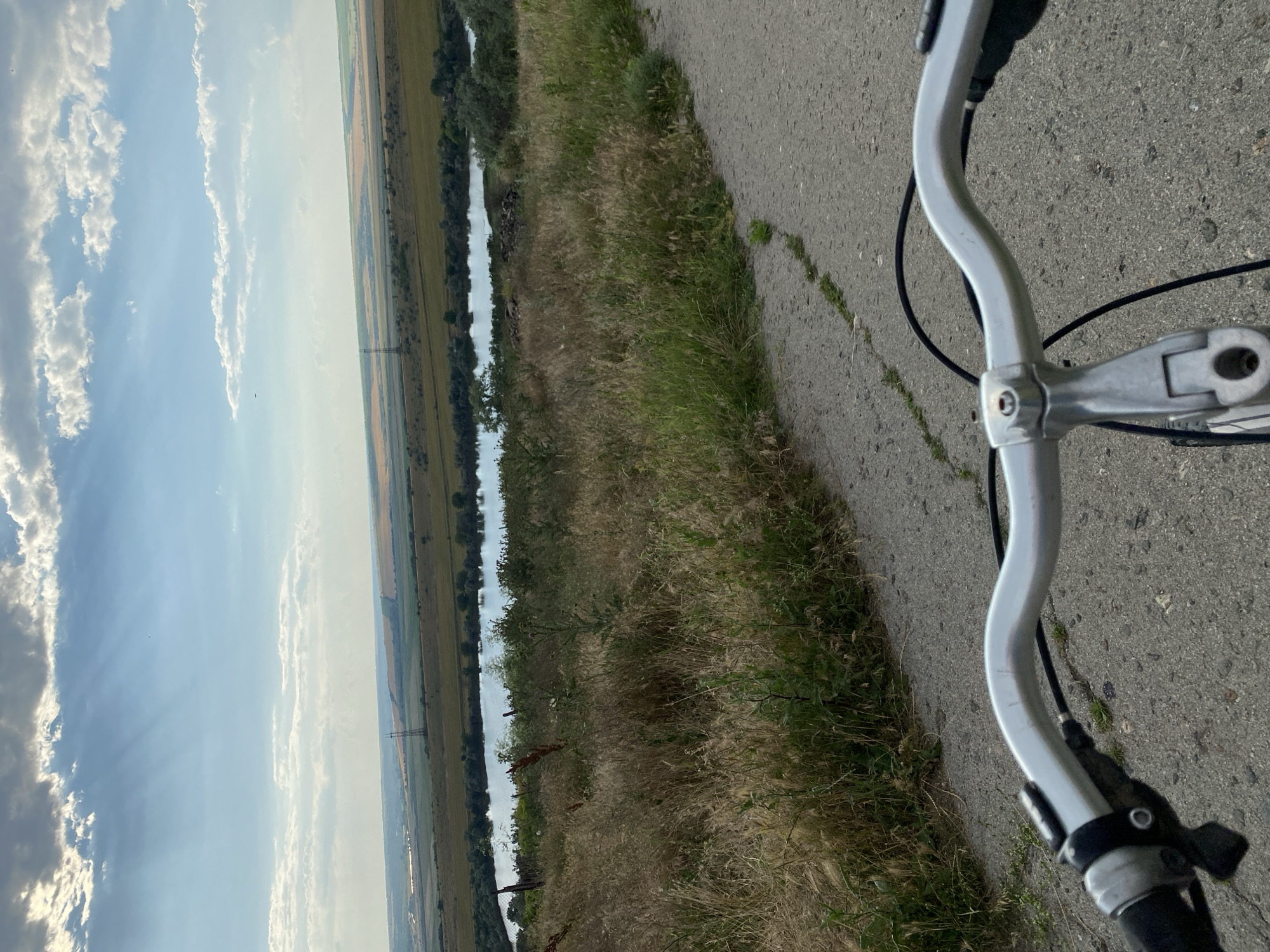 The image shows a first-person perspective of a person riding a bicycle on a paved road. The road is in the foreground, curving to the right. To the left of the road, there is tall, dry grass and some green vegetation. Beyond the vegetation, there is a body of water, likely a river or lake, with trees lining its banks. In the distance, there are rolling fields and more vegetation, with power lines and a pylon visible. The sky is partly cloudy with some rays of sunlight breaking through. The location is identified as Cairaclia, Moldova.