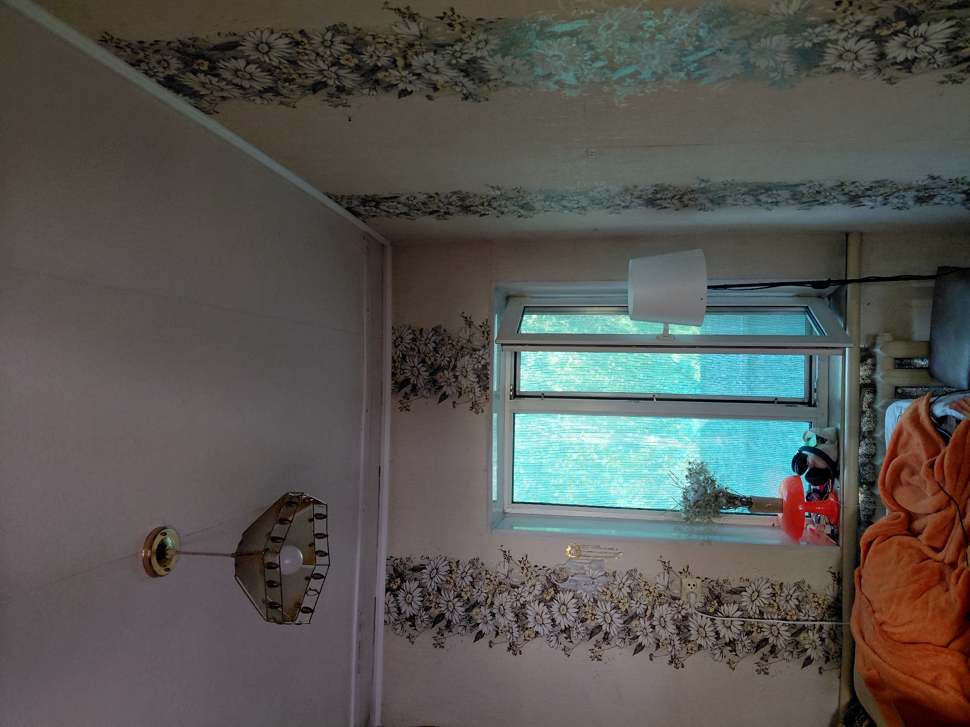 The image shows an interior view of a room with floral wallpaper. A pendant light fixture hangs from the ceiling. A window with translucent blue panes is open. On the windowsill, there is a vase of dried flowers, an orange mushroom-shaped lamp, and a bag with headphones. A white floor lamp stands to the right of the window. A bed is partially visible in the foreground, covered with an orange blanket. The walls are adorned with vertical strips of floral wallpaper featuring white and yellow daisies. The ceiling appears to be white.