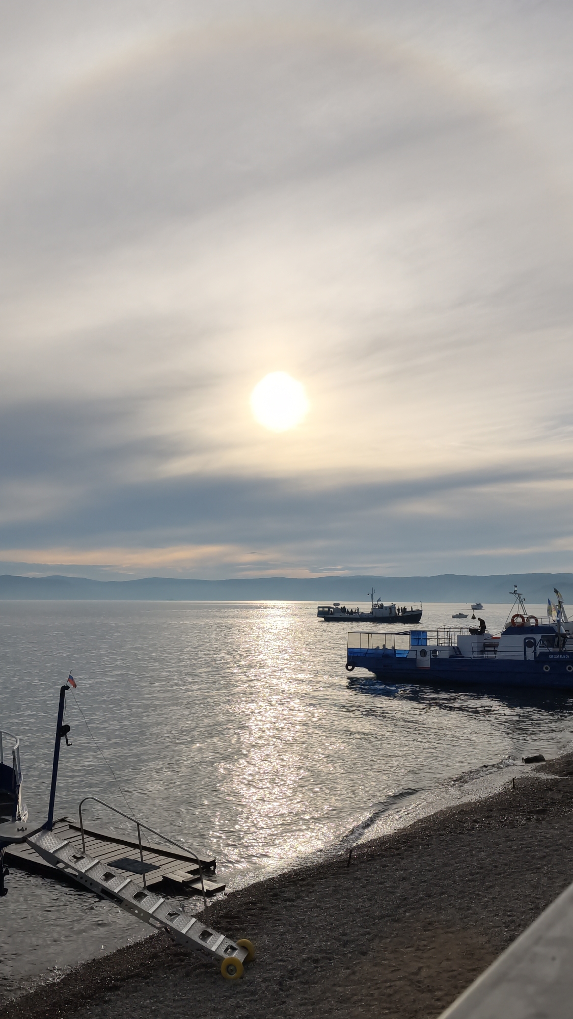 The image captures a serene scene of Lake Baikal at dusk in Listvyanka, Russia. The sun, partially obscured by clouds, casts a warm, shimmering glow on the water, creating a path of light that stretches towards the horizon. In the foreground, a small pier with a ramp extends into the calm water, where a few boats are docked. The shoreline is composed of pebbles and sand. In the middle ground, several boats are visible, some carrying passengers, navigating the lake. The distant shore is lined with rolling hills, softened by a gentle haze. The sky above is a blend of pale blues and grays, with the sun creating a subtle halo effect. The overall atmosphere is peaceful and tranquil.