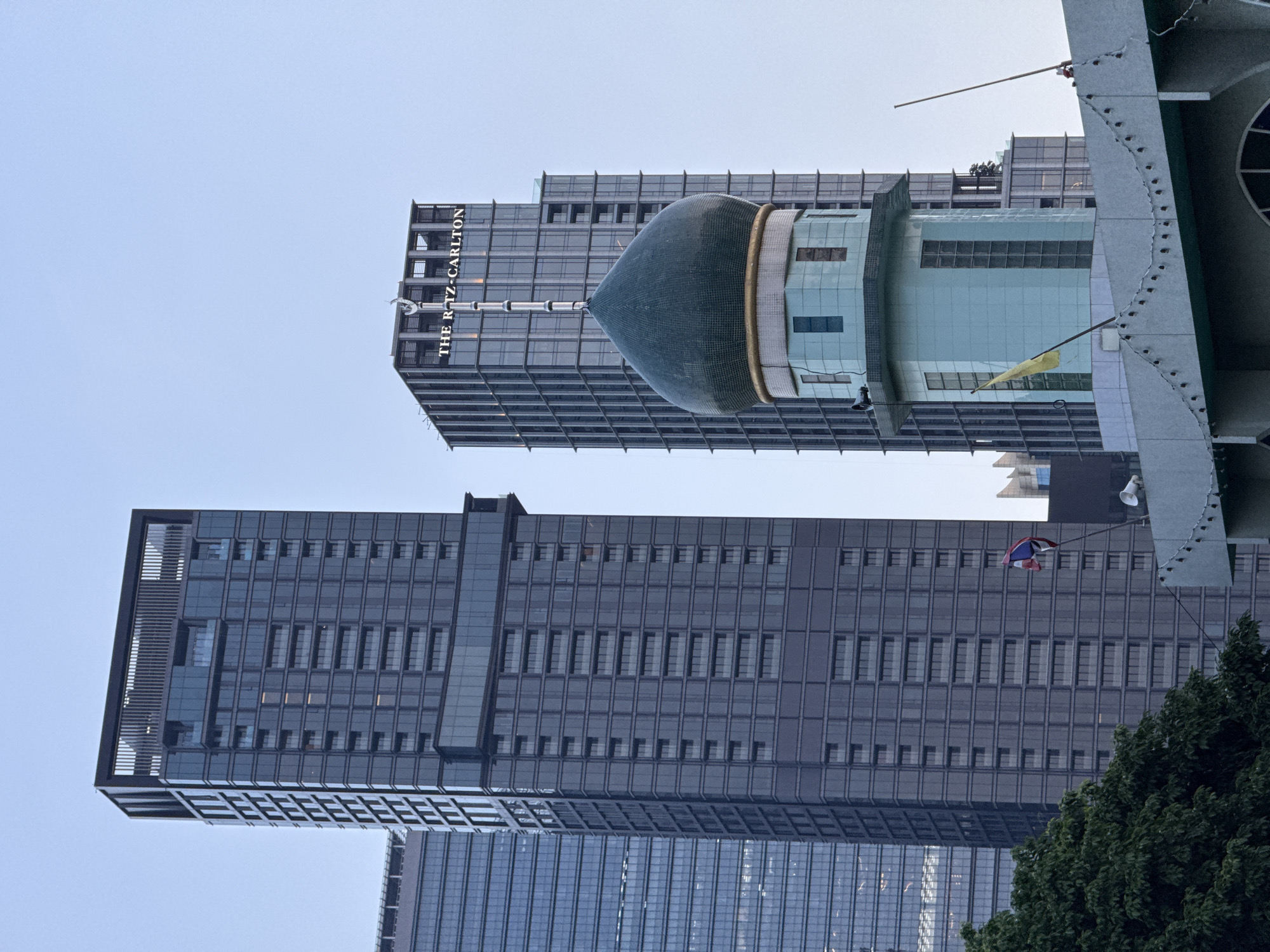 The image shows a street scene in Bangkok, Thailand, featuring a prominent mosque with a green dome and a light blue minaret, juxtaposed against modern high-rise buildings. The Ritz-Carlton hotel sign is visible on one of the buildings. A Thai flag flutters on a flagpole. Green trees frame the bottom left of the image, and a string of lights is draped along the roofline of the mosque's structure. The sky is clear and blue, suggesting it is daytime.