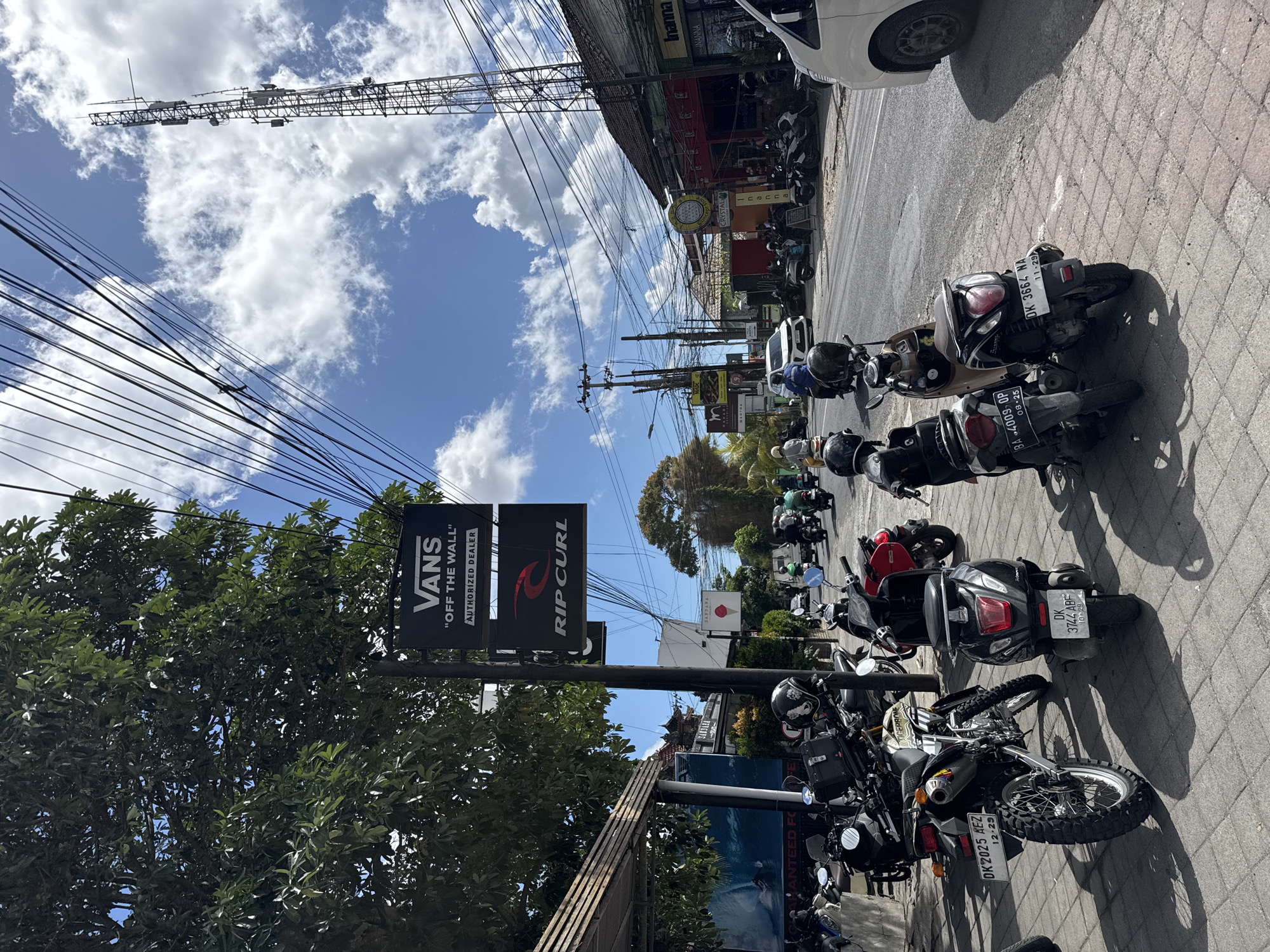 This is a daytime, eye-level, full shot of a street scene in Kuta Selatan, Indonesia. The street is lined with shops and businesses, and there are many parked motorcycles and scooters on the sidewalk. In the foreground, two scooters are parked. The scooter on the right has a black body and a white license plate that reads "DK 3664 NA 11-29." The scooter on the left is gray and has a white license plate that reads "BA 4009 OP 08-25." Behind the scooters, a motorcycle with a white and green body is parked, with a white license plate reading "DK 2025 AEZ 12-29."
A tall cell tower is visible in the background, along with a blue sky filled with white clouds. Power lines crisscross the sky. Several people are riding motorcycles and scooters on the street. A white van is parked on the right side of the road. Two black signs are hanging on a pole on the left side of the image. The upper sign reads "VANS 'OFF THE WALL' AUTHORIZED DEALER." The lower sign reads "RIP CURL." The overall impression is of a busy street scene in a tropical location.