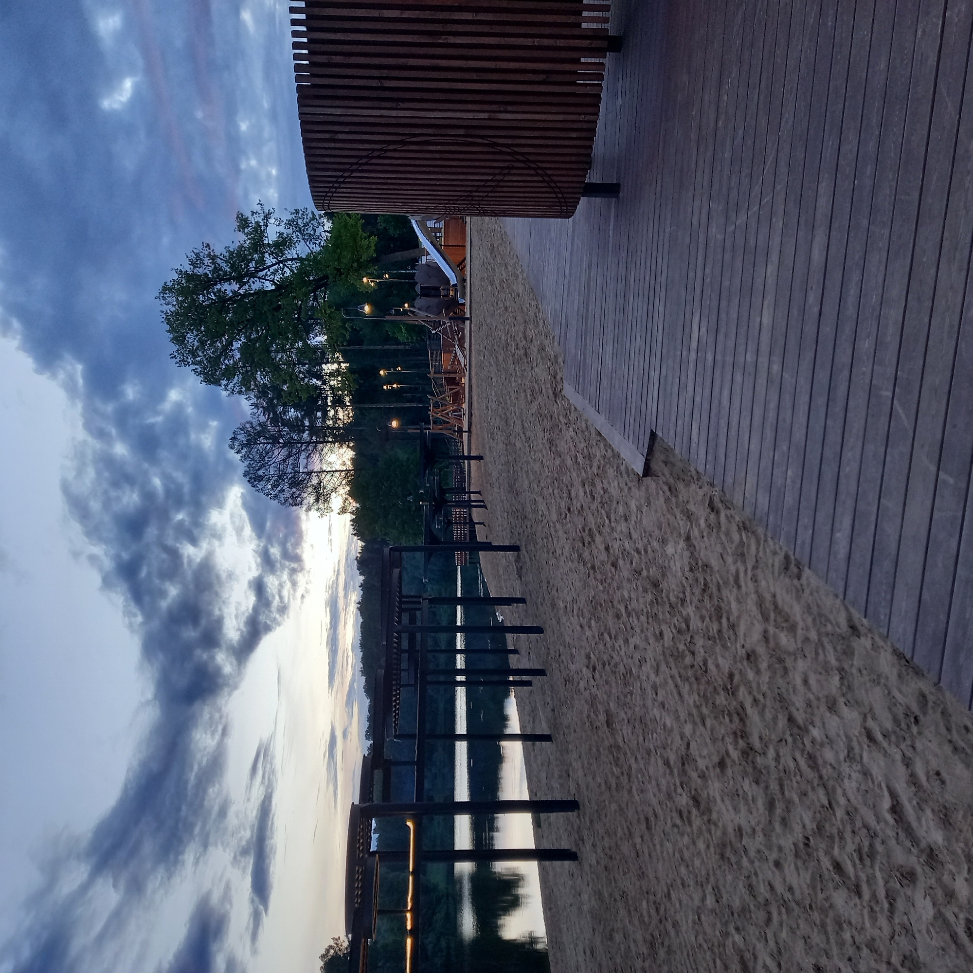 This image shows a waterfront scene in Krasnogorsk, Russia, during twilight. To the right is a wooden deck structure with vertical slats, adjacent to a sandy beach. In the distance, there's a playground and trees. The water reflects the sky, which is filled with layered clouds tinged with the colors of sunset. There are several dark post structures near the water's edge. No people are visible.