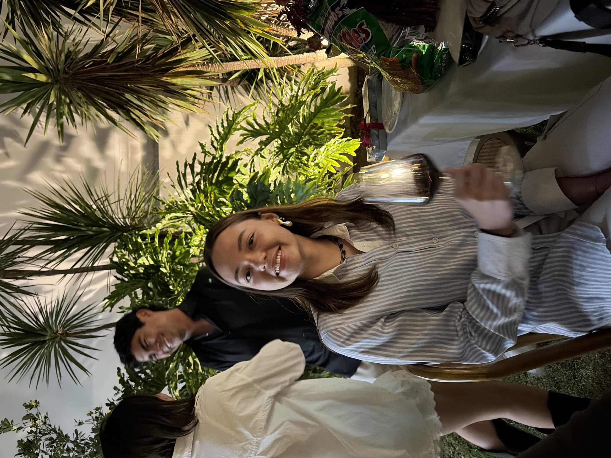The image captures an outdoor evening social gathering in an environment consistent with a private garden or patio in Puebla, Mexico. Three individuals are visible amidst lush tropical foliage under artificial illumination.
In the foreground, a young adult female with long dark brown hair is seated at a table covered with a white tablecloth. She wears a light blue and white vertical striped button-down shirt, a dark beaded necklace, and silver teardrop earrings. She holds a wine glass containing dark red liquid in her right hand and is smiling directly at the camera. Her lower body is partially visible in light-colored trousers.
To her left, a second young adult female is seen from the back, her head and upper torso visible. She has dark hair and wears a loose white button-down shirt, with bare legs and dark boots partially visible. She is facing towards the third individual, suggesting an interaction.
The third individual, a young adult male with dark hair, stands in the mid-ground between the two females. He wears a black collared shirt and is smiling towards the female in white.
The background features dense, vibrant green tropical or subtropical plants, including large-leafed varieties and tall, narrow-leafed plants, likely illuminated from below, casting shadows onto a light-colored wall. The ground beneath the individuals appears to be grass or artificial turf.
On the white-clothed table to the right of the seated female, a white round plate, a green and white branded bag of potato chips identified as "Sabritas" by visible text and color scheme, two rectangular gift boxes wrapped with red ribbons, and a light-colored shoulder bag with a dark strap are present.
