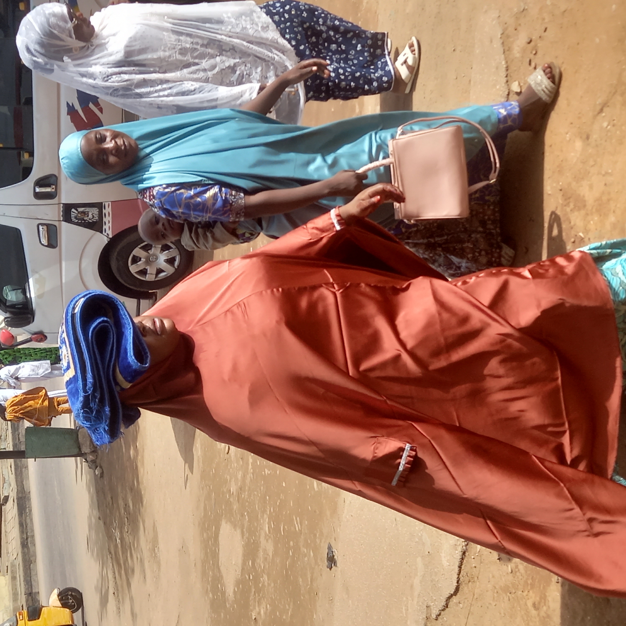 Three women dressed in traditional Islamic clothing stand near a white van on what appears to be a street in Dadi Riba, Nigeria. The woman closest to the camera wears an orange robe with a blue head covering. Another woman wears a light blue robe and carries a pink purse. A child is nestled in the light blue-clad woman's arm. The third woman wears a white robe and white sandals with a dark blue floral skirt. The ground is dirt or sand. In the background, there's a street with a yellow vehicle and a light post.