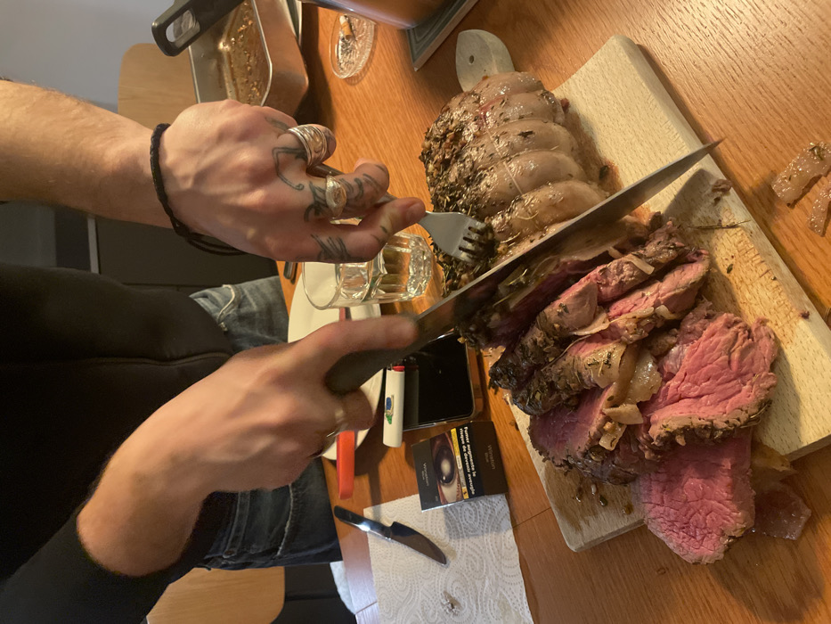 A medium-rare beef roast is being sliced on a wooden cutting board by a person wearing a black shirt and jeans. The roast is surrounded by onions and herbs. A fork is used to stabilize the roast as it is sliced. A pack of cigarettes, a lighter, a cellphone, and a knife rest on a paper towel nearby. The person has several rings and tattoos on their hands. Dishes, including a pan with liquid, are visible in the background on a wooden table.