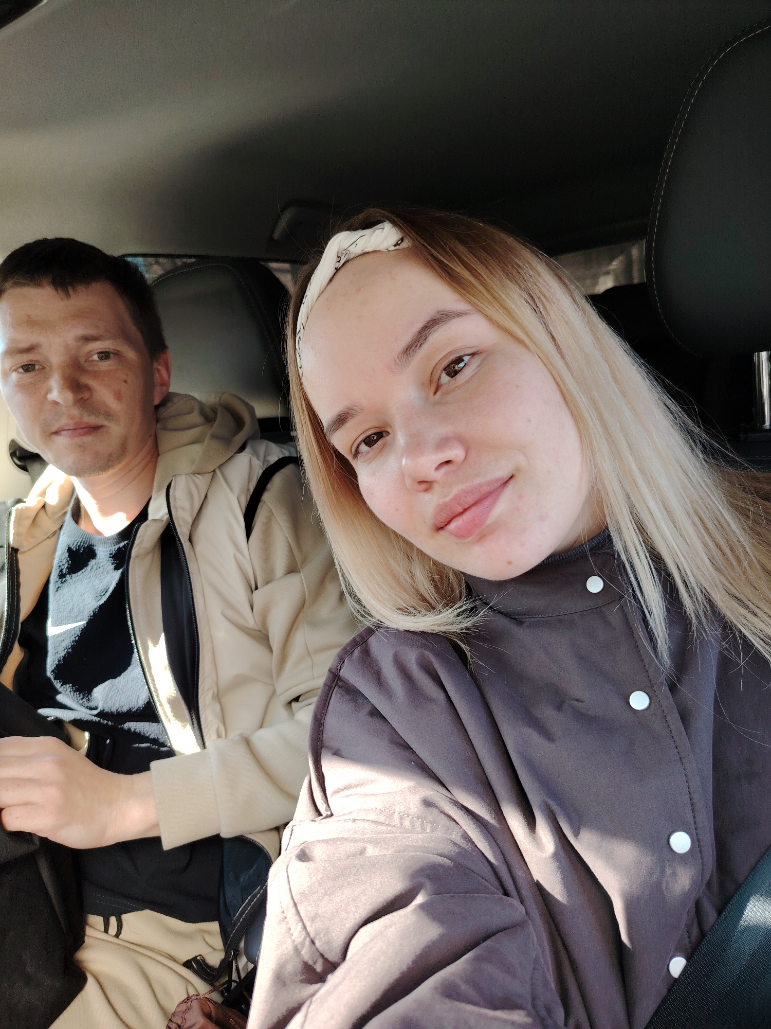 Two individuals are inside a car. A man with short brown hair is on the left, wearing a light brown jacket over a black and white shirt. A woman with blonde hair and a white headband is on the right, wearing a brown jacket with white buttons and a black seatbelt. The interior of the car, including the black headrest, is visible. The location is Chaikovsky, Russia.