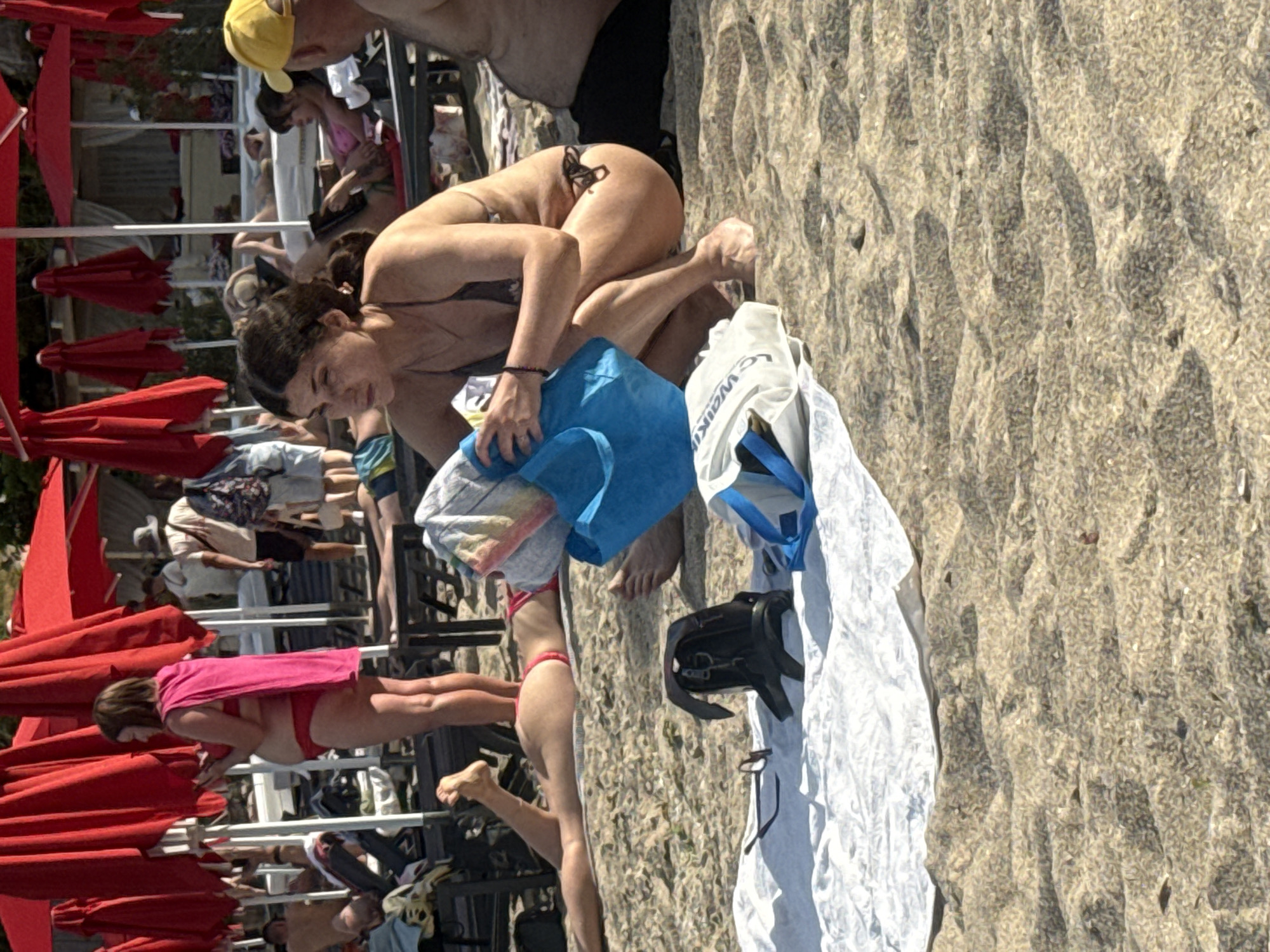 In Odesa, Ukraine, on a sandy beach, a woman with dark hair, wearing a dark bikini, is crouched down. She is holding a light-colored towel in her hands and has a bright blue bag next to her. A white towel and a black backpack are on the sand in front of her. In the background, several people are visible, some relaxing on lounge chairs under red umbrellas. The overall scene depicts a typical day at a crowded beach.