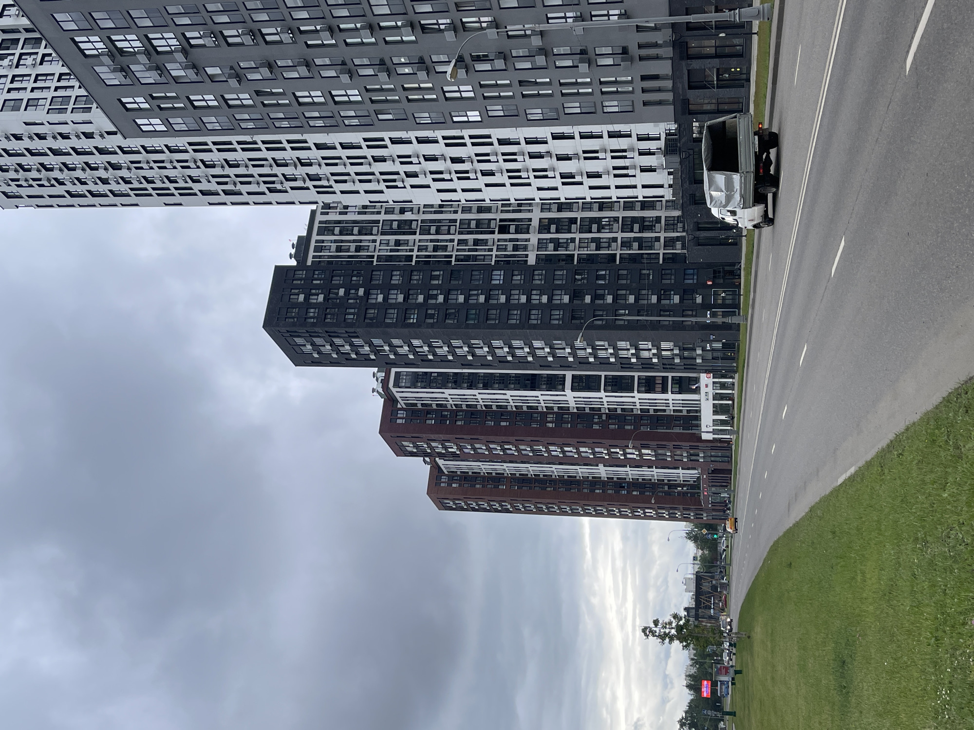 The image depicts a wide, grey asphalt road in Moscow, Russia, with multiple lanes of traffic. The road is flanked by a lush green grass verge on the left and a row of tall, modern apartment buildings on the right. These buildings are predominantly dark grey and white, with numerous windows and balconies. Several vehicles are visible on the road, including a white truck with a covered cargo bed moving away from the camera. In the distance, there are other cars and what appear to be pedestrian crossings and traffic signals. The sky is overcast with heavy, grey clouds, suggesting an impending or recent rainfall. The overall scene conveys a sense of urban development and daily life in a Moscow residential area.