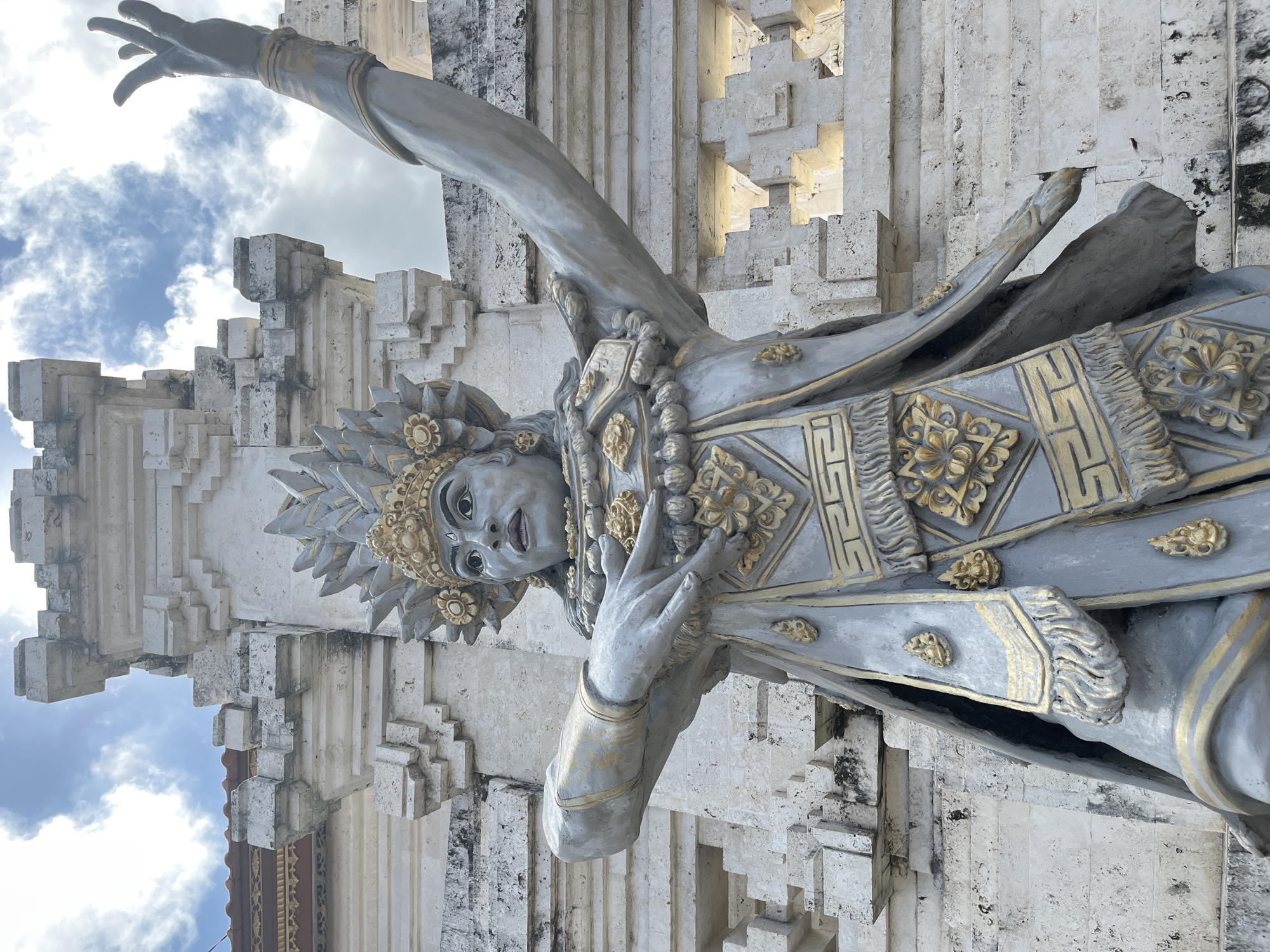 A full-shot, low-angle photograph captures a statue of a Balinese dancer in traditional attire, standing in front of a weathered stone temple structure. The statue is predominantly gray with gold accents, adorned with intricate patterns on its headdress, costume, and jewelry. Its arms are posed in a dance-like gesture, with one hand pointing upwards and the other extended outwards. The temple, made of light-colored stone with a textured surface, features layered architectural elements and shows signs of age, with some discoloration. The background reveals a cloudy blue sky. The image is set in Kuta Selatan, Indonesia, likely at a cultural or religious site.