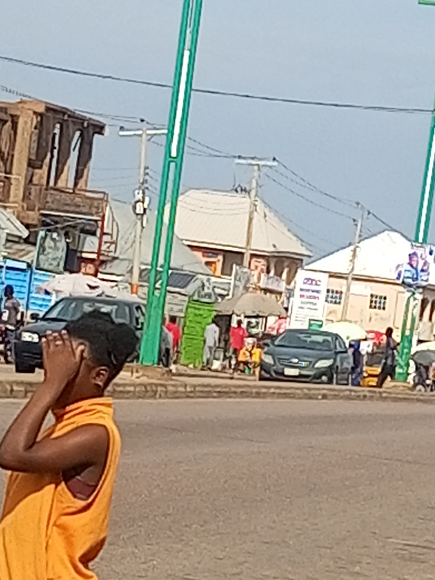 The image shows a street scene in a city. In the foreground, a person is partially visible from the chest up, wearing an orange sleeveless shirt and covering their face with their hands. The person appears to be looking down and to the left.
In the background, there are several buildings of various sizes and styles. Some appear to be shops with awnings and signage. A blue truck is parked on the left side of the street. Several cars, including a dark-colored sedan in the center, are also present. Various people are walking on the sidewalk and near the vehicles. There are also some stalls or temporary structures set up on the side of the road, possibly for vendors. Power lines and utility poles are visible against the sky. The overall environment suggests a busy urban area with a mix of commercial and residential buildings. The lighting indicates it is daytime.