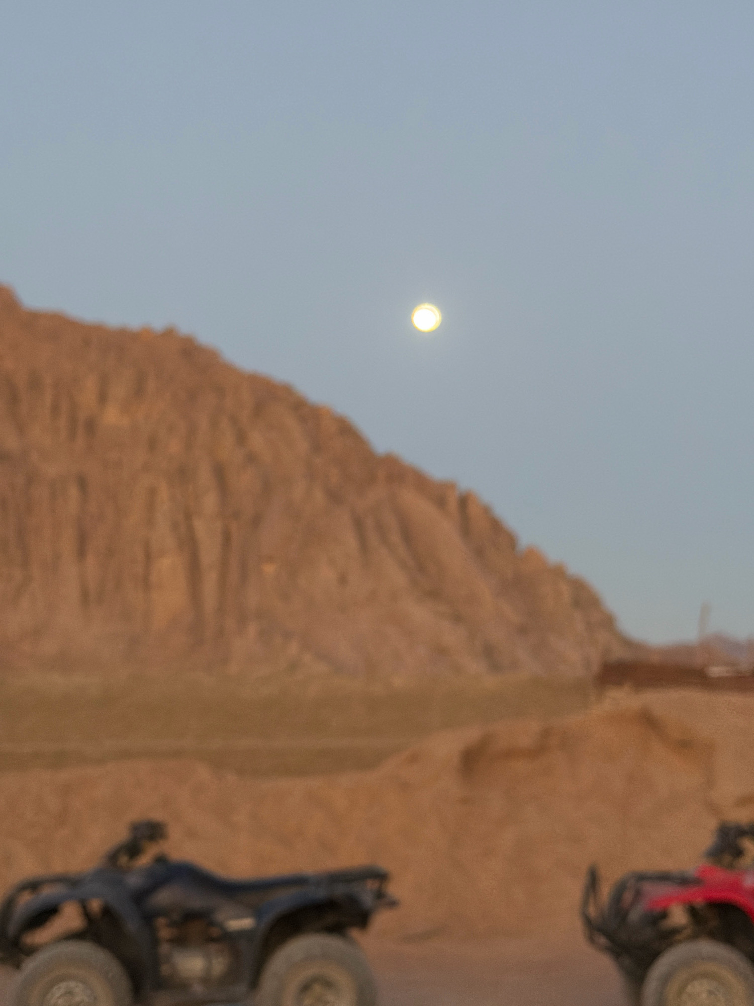 The image depicts a desert landscape in Sharm El Sheikh, Egypt, under a pale blue sky. A bright, full moon is visible high in the sky. In the foreground, two all-terrain vehicles (ATVs) are parked. One ATV is black and is positioned on the left side of the frame, showing its front and side. The other ATV is red and is visible on the right side of the frame, showing its rear and side. The background features a large, barren, rocky mountain range with distinct striations, suggesting a geological formation shaped by erosion. The overall atmosphere is serene and evokes the natural beauty of the desert at dusk or dawn.