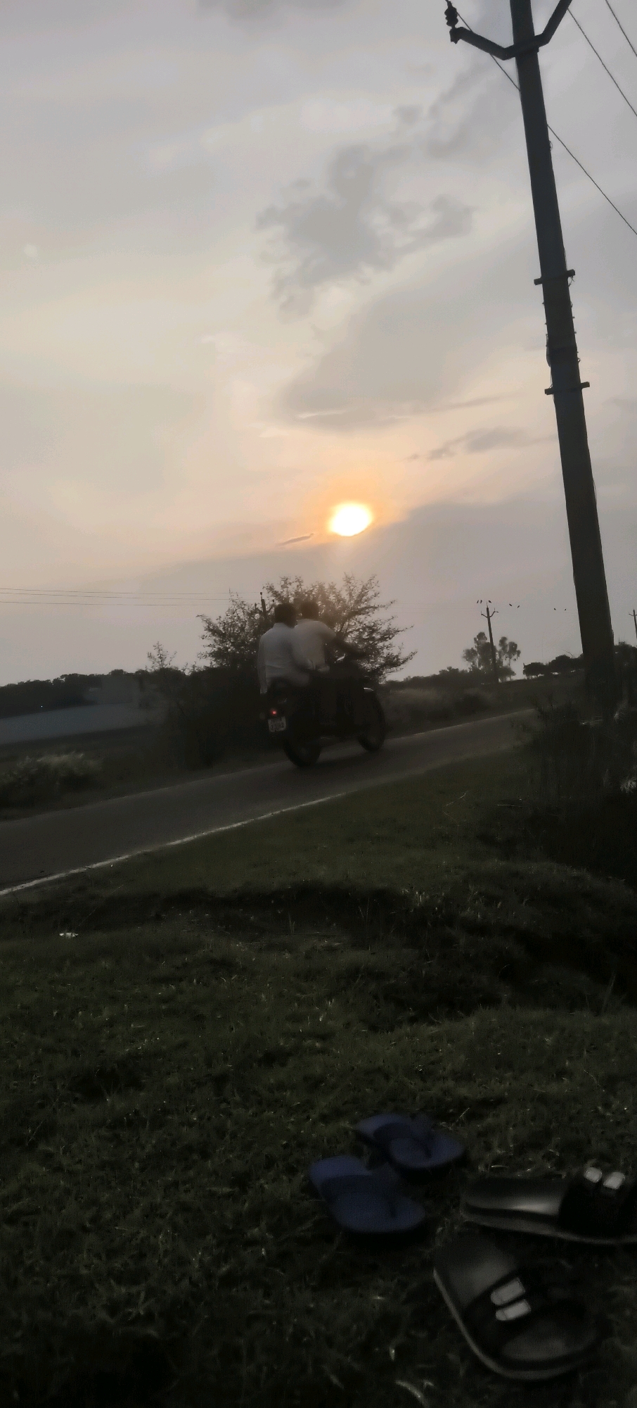 In Narayanpur, India, the image captures a tranquil rural scene at dusk. The sun, a soft orb of light, is descending in a hazy sky, casting a warm glow over the landscape. A solitary road cuts through a grassy expanse, flanked by lush greenery. On the road, a motorcycle carries two individuals, their figures silhouetted against the fading light. The scene is framed by a tall utility pole on the right, its wires stretching across the sky, and a cluster of trees in the middle ground. In the foreground, a pair of blue flip-flops rests on the grass, adding a touch of everyday life to the serene setting. The overall atmosphere is one of peace and quietude, typical of rural India during the twilight hours.