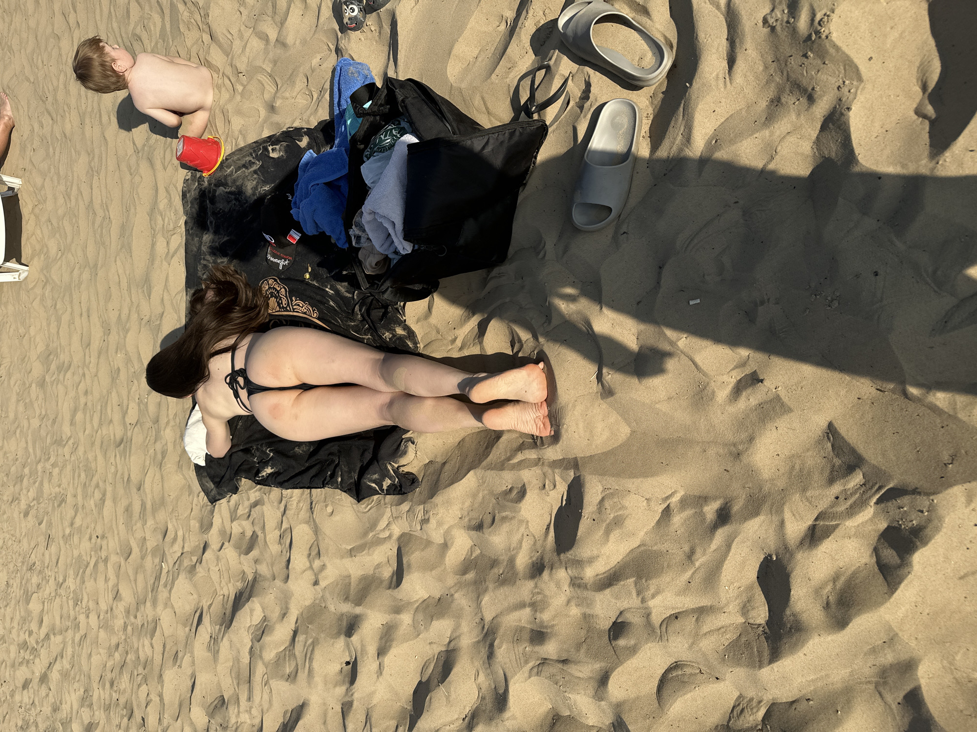 The image depicts a sandy beach, likely in Matiushino, Russia, based on the request. A woman in a black bikini lies on a black towel, with her legs extended away from the viewer. Next to her is a black bag filled with clothing and other items. A young child lies shirtless in the sand, with a red bucket nearby. A pair of gray slides are also visible on the sand near the bag. The scene appears to be captured from an elevated perspective, with shadows cast across the sand.