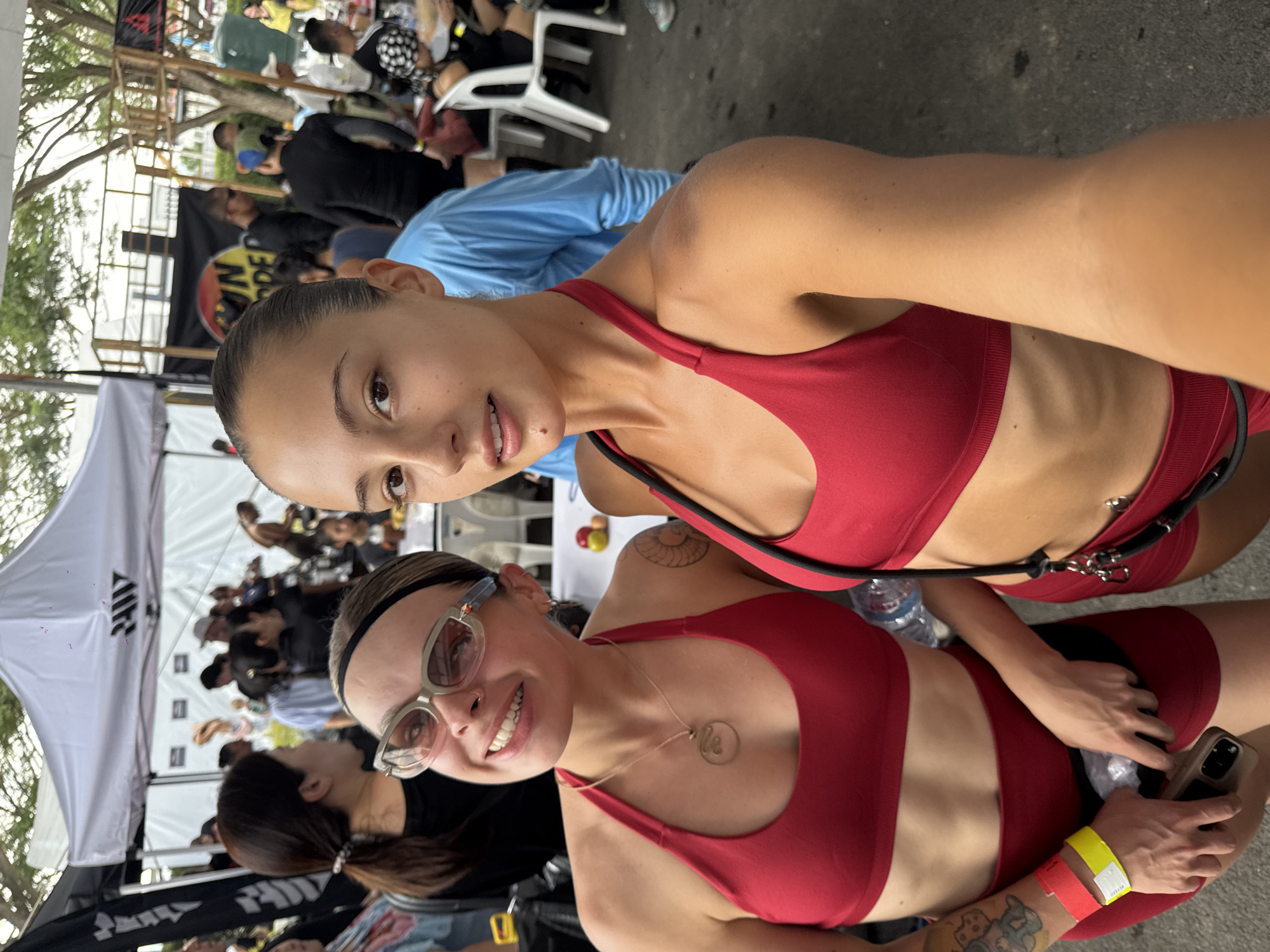 The image shows two women wearing matching red sports bras and shorts, with a crowd of people in the background. The women are close together, likely taking a selfie. The woman in the foreground is wearing sunglasses and a necklace. The woman in the background has a black harness-like strap across her torso. A water bottle is visible between them. The crowd behind them appears to be gathered at an event with tents and banners. The image was likely taken outdoors, based on the lighting and surrounding foliage. Given the location as บ้านสำนักตะแบก, Thailand, the crowd may be attending a local event.