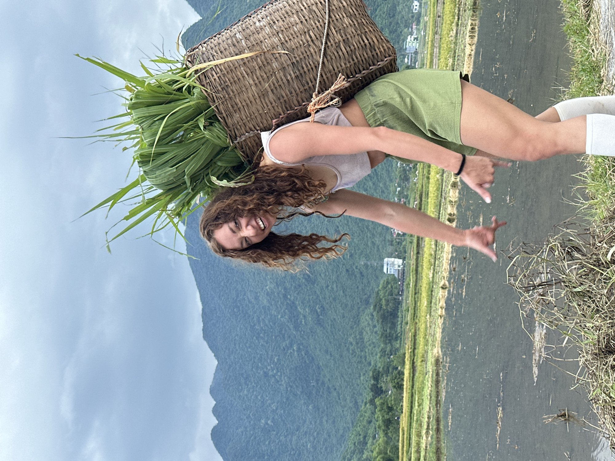 In Chiềng Châu, Vietnam, a woman with curly brown hair, wearing a light pink crop top and green shorts, is captured mid-stride. She is carrying a large, woven basket on her back, filled with lush green foliage. Her mouth is open in a wide smile, and her right arm is extended outwards, fingers splayed, as if gesturing or reaching. Her left arm is bent at the elbow, also extended forward. She is wearing white knee-high socks. The background features a vast expanse of flooded rice paddies, with verdant, rolling hills and mountains in the distance. The sky is overcast with a gradient of light and dark clouds.