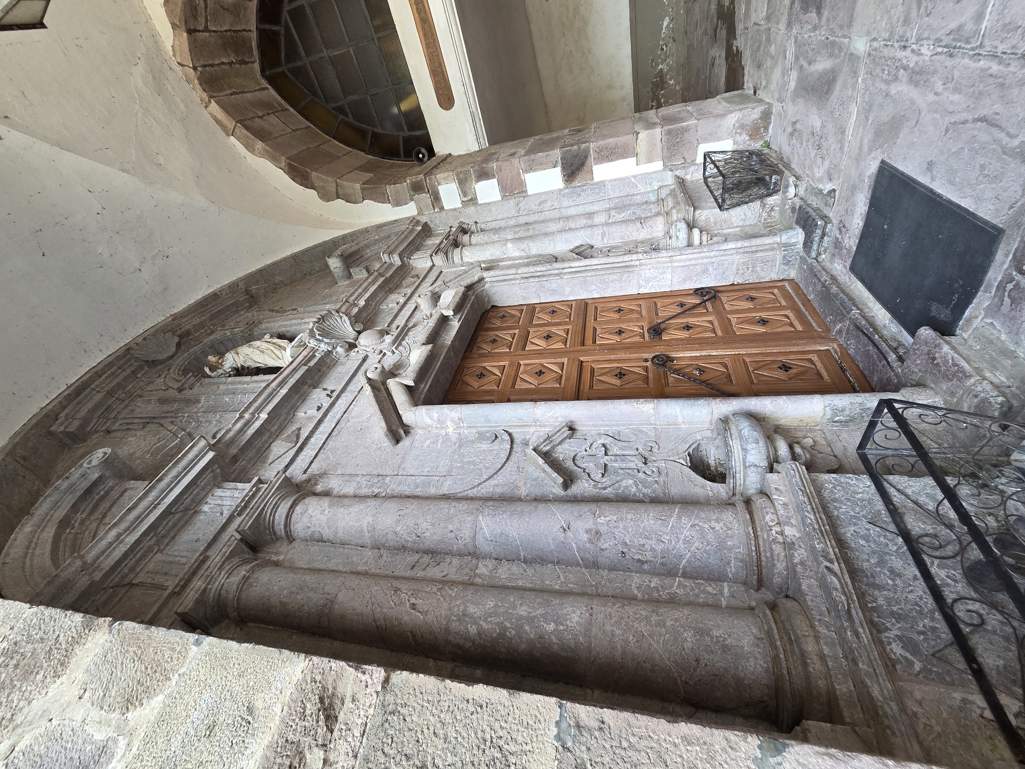 The image shows a stone facade with a wooden door. To the left of the door, there are two fluted columns and a small carved cross with a basin underneath. Above the door, there is a statue of a figure in a niche. To the right of the door, there is a stone pillar and an arched passageway with a window. The ground in front of the door is paved with stone. A black doormat is placed in front of the door. A metal grate and a small metal basket are also visible on the ground. The overall scene is of an old building's entrance, possibly a church or historical structure. The location is identified as the city of Baztan, Spain. There are no people visible in the image.