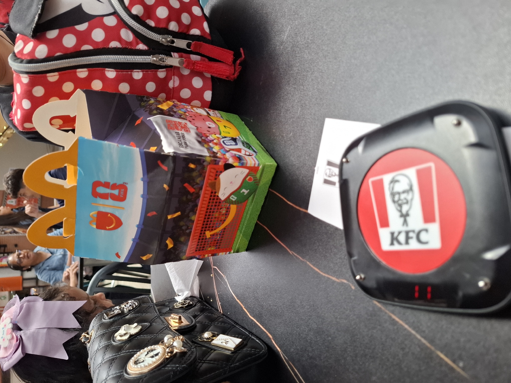 In an indoor setting, possibly a restaurant or dining area in Pampatar, Venezuela, the image captures a collection of items on a dark, textured surface. A red and white polka dot bag with silver zippers is prominent in the upper left. Next to it, a colorful McDonald's Happy Meal box is visible, featuring cartoon characters and a stadium scene. To the right, a black pager with the KFC logo is displayed, showing red indicator lights at the bottom.
In the background, several individuals are partially visible. One person in a blue shirt is looking towards the camera, while another person is obscured by the objects in the foreground. A black quilted handbag adorned with various charms and a light purple bow is in the lower right portion of the image. The overall impression is of a casual gathering or mealtime. The lighting suggests it is daytime.
