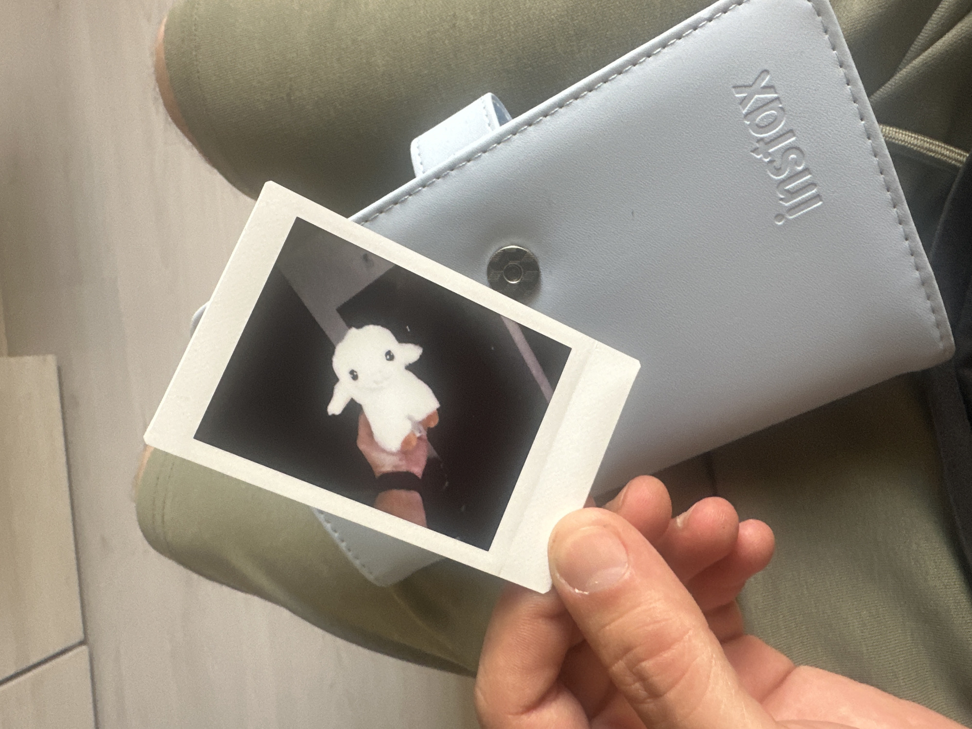 A person is holding a Polaroid photograph in their hand. The photograph depicts a small, white, plush toy. The person is sitting, with their lap visible in the background, and a light blue Instax camera case is resting on their lap. The photograph is the primary focus, held between the thumb and index finger of the person's right hand. The background is slightly out of focus. The photograph itself shows a dark, indoor setting with the plush toy being held up in someone's hand. The presence of the Instax camera case implies that the photograph was likely taken with an Instax camera. There is no information to determine the location as Slavyani, Bulgaria.