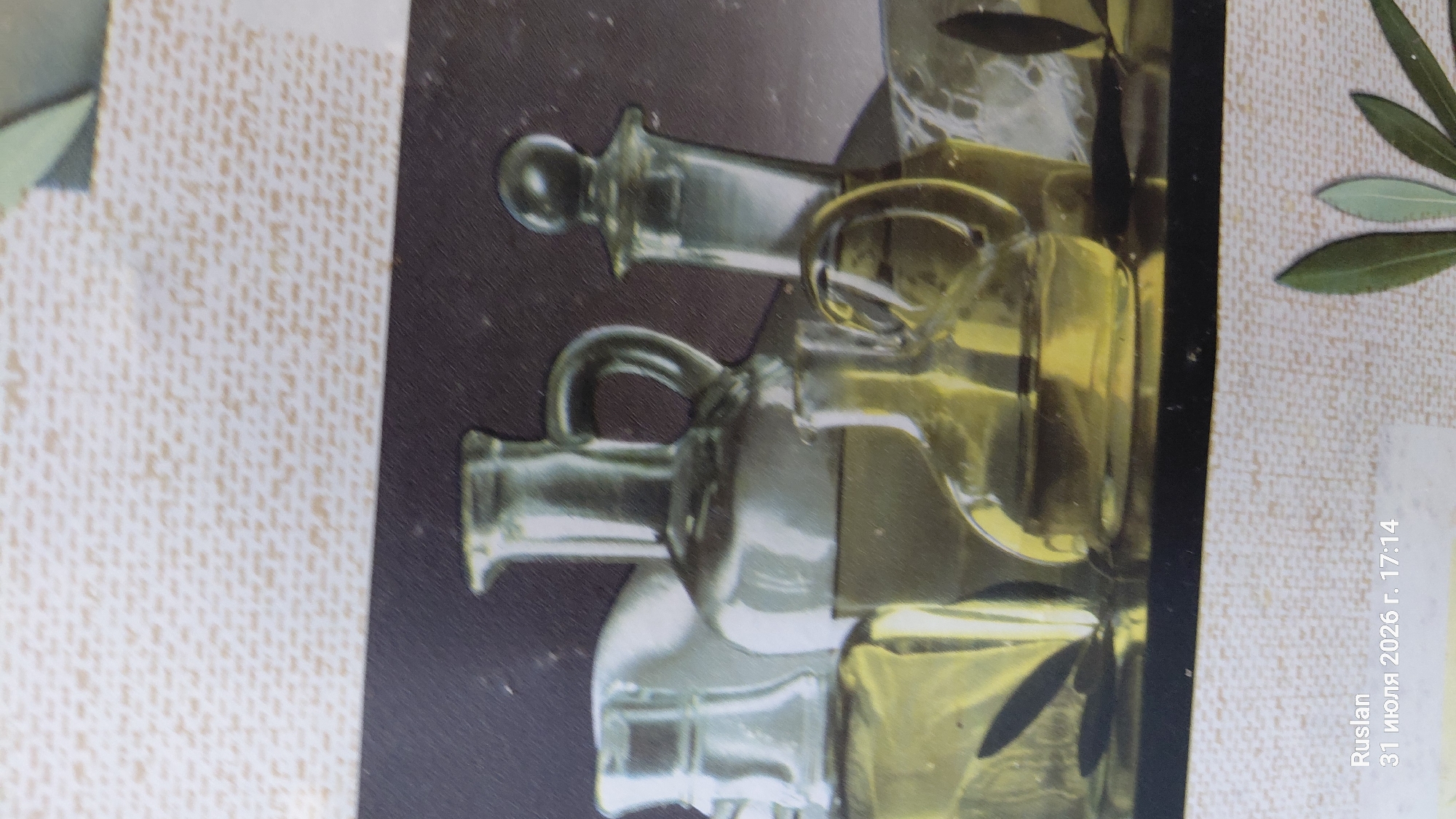 The image shows a still life composition of several glass bottles filled with a yellow liquid, presumably olive oil, against a dark background with a textured overlay. The bottles vary in size and shape, with some featuring handles and stoppers. The lighting highlights the translucence of the glass and the color of the liquid. In the lower left corner, there is text that reads "Ruslan 31 июля 2026 г. 17:14", which is a watermark indicating a name and a date and time. There is no indication of the city of Oleksandriia, Ukraine, in the image.