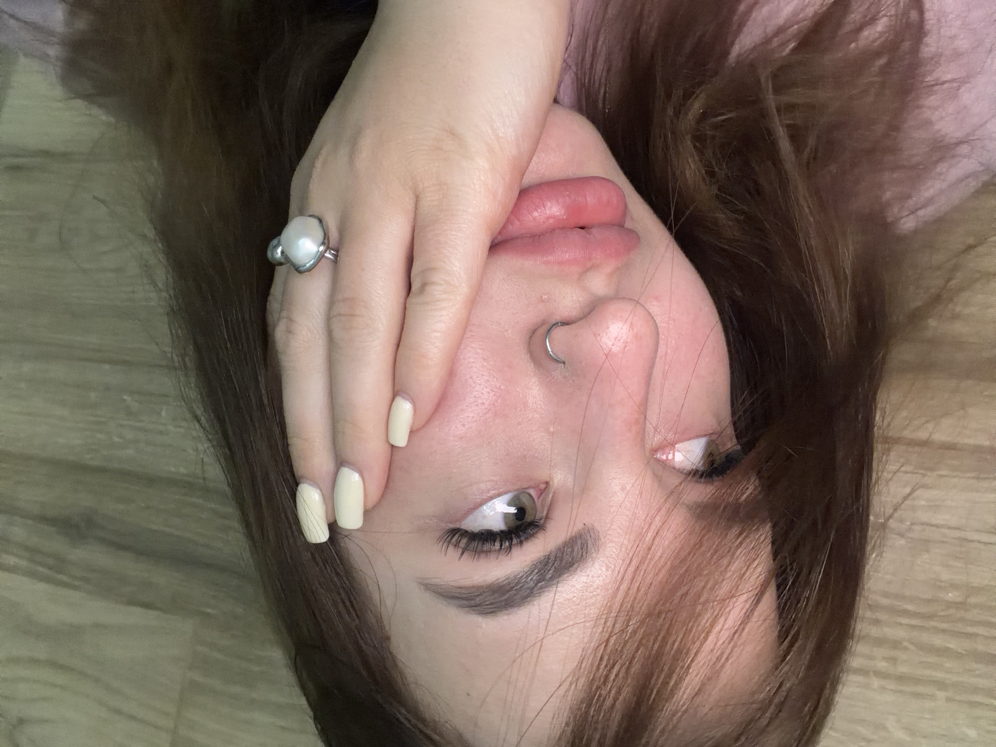 An overhead close-up shot captures a person lying on their back on a wooden floor in an indoor setting in Krasnodar, Russia. The person's head is tilted, with their light brown hair fanned out around them. One hand is partially covering their mouth and nose.
The primary subject is the person's face, visible from the forehead down to the chin, obscured partially by the hand. Their skin is fair. They have light-colored eyes, appearing greenish-hazel, with long, dark eyelashes. Their eyebrows are well-defined and dark. A small silver hoop nose piercing is visible on their right nostril. Their lips are slightly parted and appear naturally full.
The hand covering part of the face has short, square-shaped nails painted in a light beige or off-white color. A prominent silver-toned ring with a large, irregular-shaped white pearl is worn on the index finger of this hand. The surrounding hair is medium brown with some lighter highlights, appearing soft and slightly wavy. The background is a light-colored wooden floor with visible grain.
The activity appears to be a posed close-up portrait or selfie while lying down. No specific emotions are clearly discernible due to the partial facial obstruction and the pose, but the overall impression is contemplative or relaxed. There is no visible text in the image.