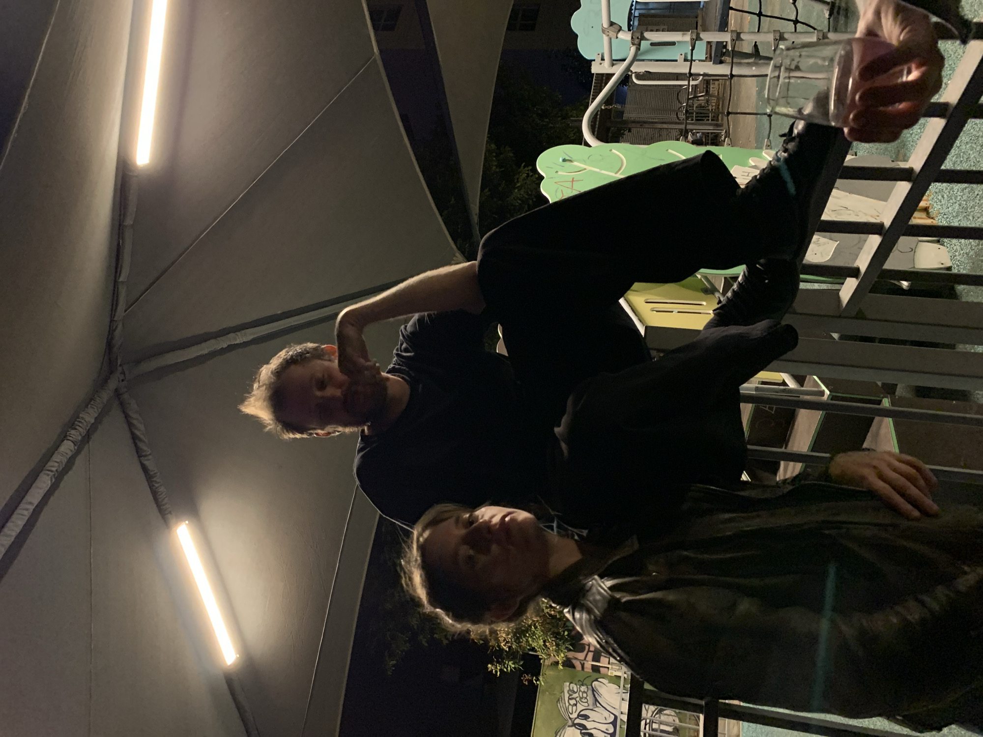 The image shows three people at night in what appears to be a covered outdoor area, possibly a playground. Two individuals are visible towards the lower portion of the frame. One person is wearing a dark jacket, while the other appears to have on a dark t-shirt. The third person is positioned at the top of the frame. They are holding a glass. Fluorescent lights are present in the background. Based on the context clues in the images, the location might be in Tel Aviv, Israel, as this location is mentioned in the original request.