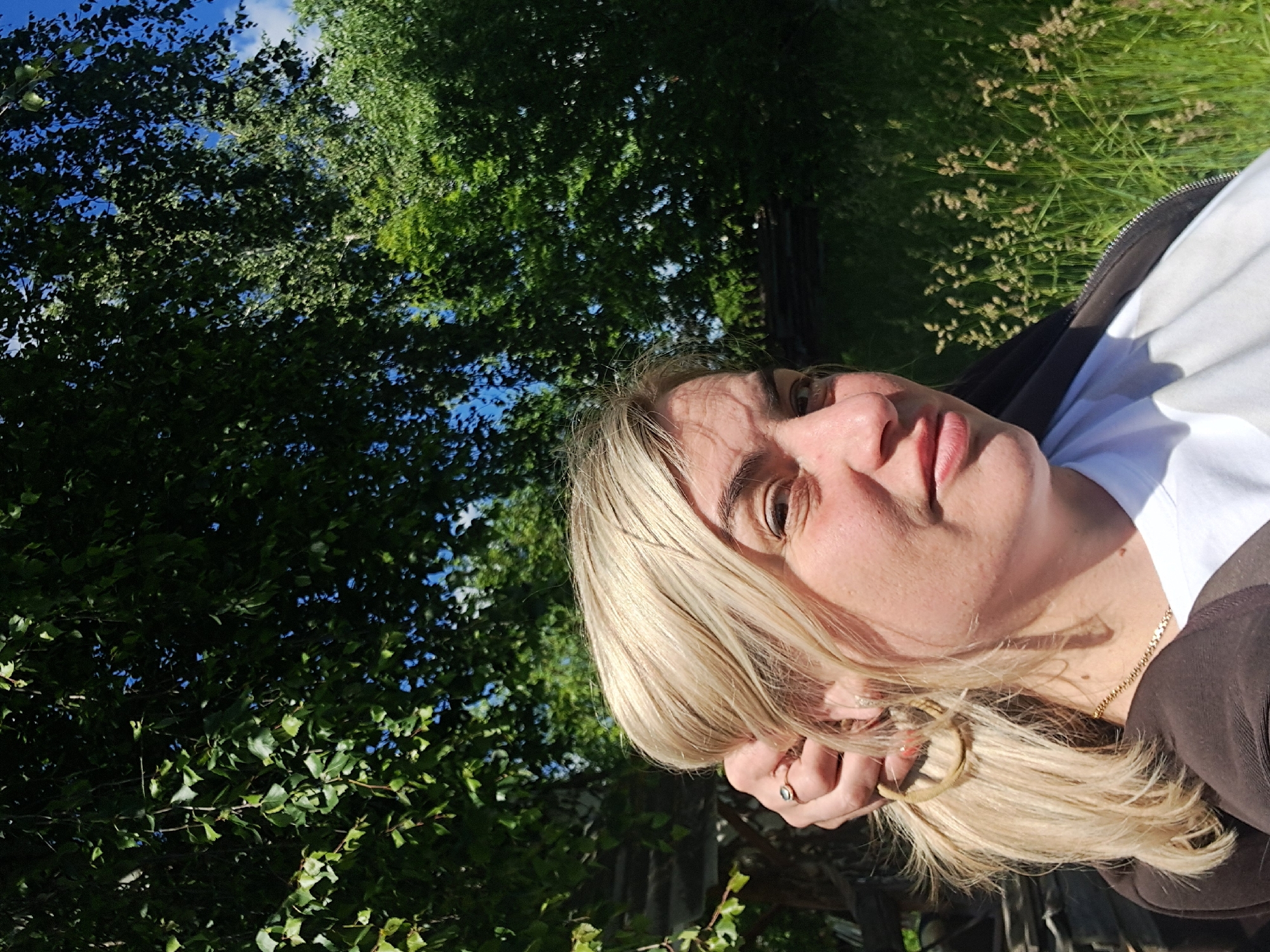 A bright, sunlit, outdoor portrait features a fair-skinned woman with blonde hair, visible from her upper chest to the top of her head. She is looking directly at the camera with a neutral to slightly smiling expression. Her blonde hair is styled loosely, with some strands falling around her face, and one hand is raised, possibly touching her hair or face. She wears a white t-shirt under a dark, possibly brown or black, zippered jacket. A delicate gold chain necklace is visible around her neck, and a ring is on her finger.
The background is dominated by dense green foliage of trees, suggesting a natural setting like a park or forest. Patches of clear blue sky with some white clouds are visible through the gaps in the leaves. To the right side of the image, behind the woman, there's an area of tall, light brown and green grass or reeds, indicating a wilder or less manicured landscape. The strong sunlight illuminates the woman's face and hair, creating highlights and casting some gentle shadows, suggesting it is daytime, likely morning or late afternoon. No text is visible in the image. The scene is set in an outdoor, rural or natural area.