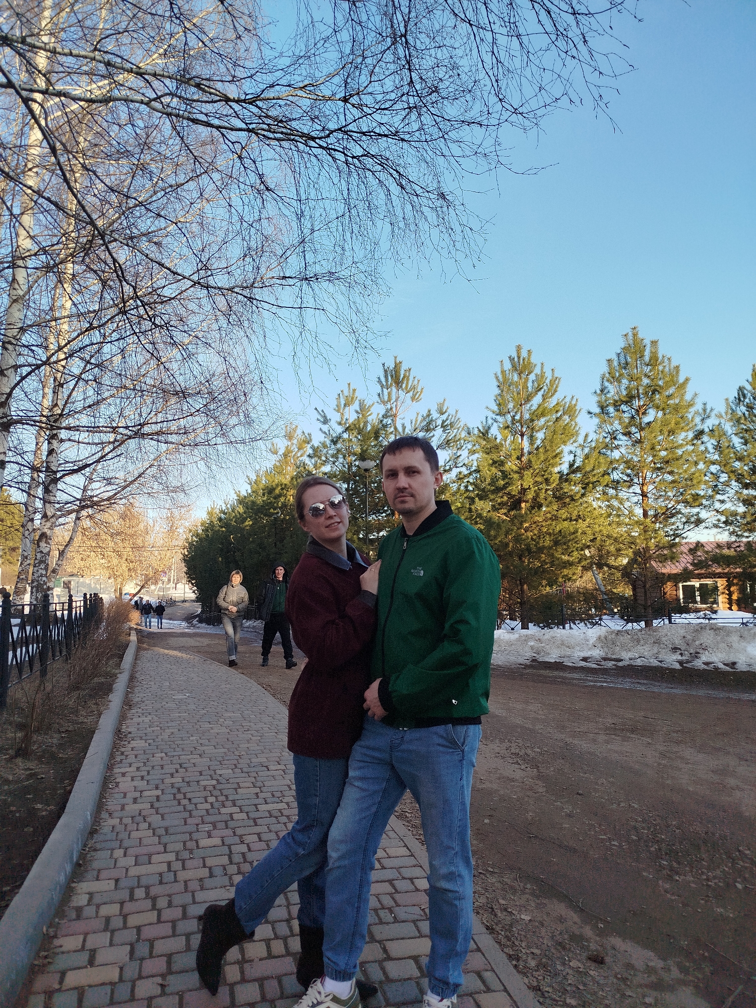 A couple, a woman and a man, stand embraced on a brick pathway. The woman wears a maroon sweater, blue jeans, and sunglasses. Her right leg is extended outward. The man wears a green jacket and blue jeans. Behind them are pine trees and remnants of snow. To the left of the couple is a metal fence bordering the pathway, along with bare trees. In the background, two figures walk along the pathway. Further in the background is a building with a brown roof. The location is Izhevsk, Russia.