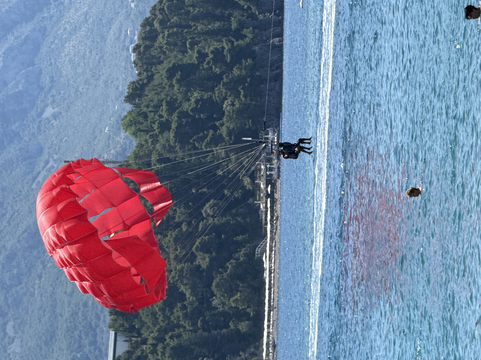 This image captures a scenic view of parasailing activity in the waters near Budva, Montenegro. In the foreground, the bright red canopy of a parasail dominates the upper left portion of the frame, its fabric billowing in the wind. Below the parasail, two individuals are suspended in the air, seated in a harness, enjoying the experience of parasailing. They appear to be moving across the turquoise water, leaving a white wake behind them.
In the mid-ground, the clear blue water stretches out, reflecting the sunlight. A person is visible swimming or wading in the water, their head and shoulders above the surface. Further in the distance, along the coastline, a strip of beach is lined with numerous white lounge chairs and umbrellas, suggesting a popular tourist area. Beyond the beach, lush green trees cover the slopes of a mountainous landscape, with hints of buildings nestled within the foliage. The overall scene conveys a sense of leisure and outdoor recreation in a beautiful coastal environment.