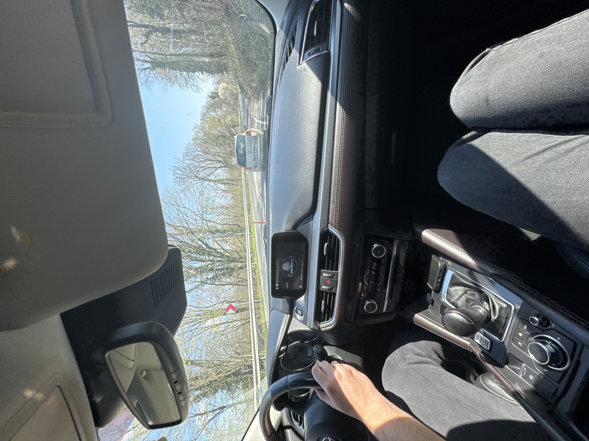 The image depicts the interior of a car, a Mazda CX-5, driving on a road with trees lining the sides, likely in Staro Oriahovo, Bulgaria, based on the license plate of the car in front (presumably Bulgarian). The dashboard is visible, displaying information on the car's infotainment screen. The driver's hand is visible on the steering wheel, and the driver is wearing dark colored pants. The sun is shining. The interior of the car is partly leather.