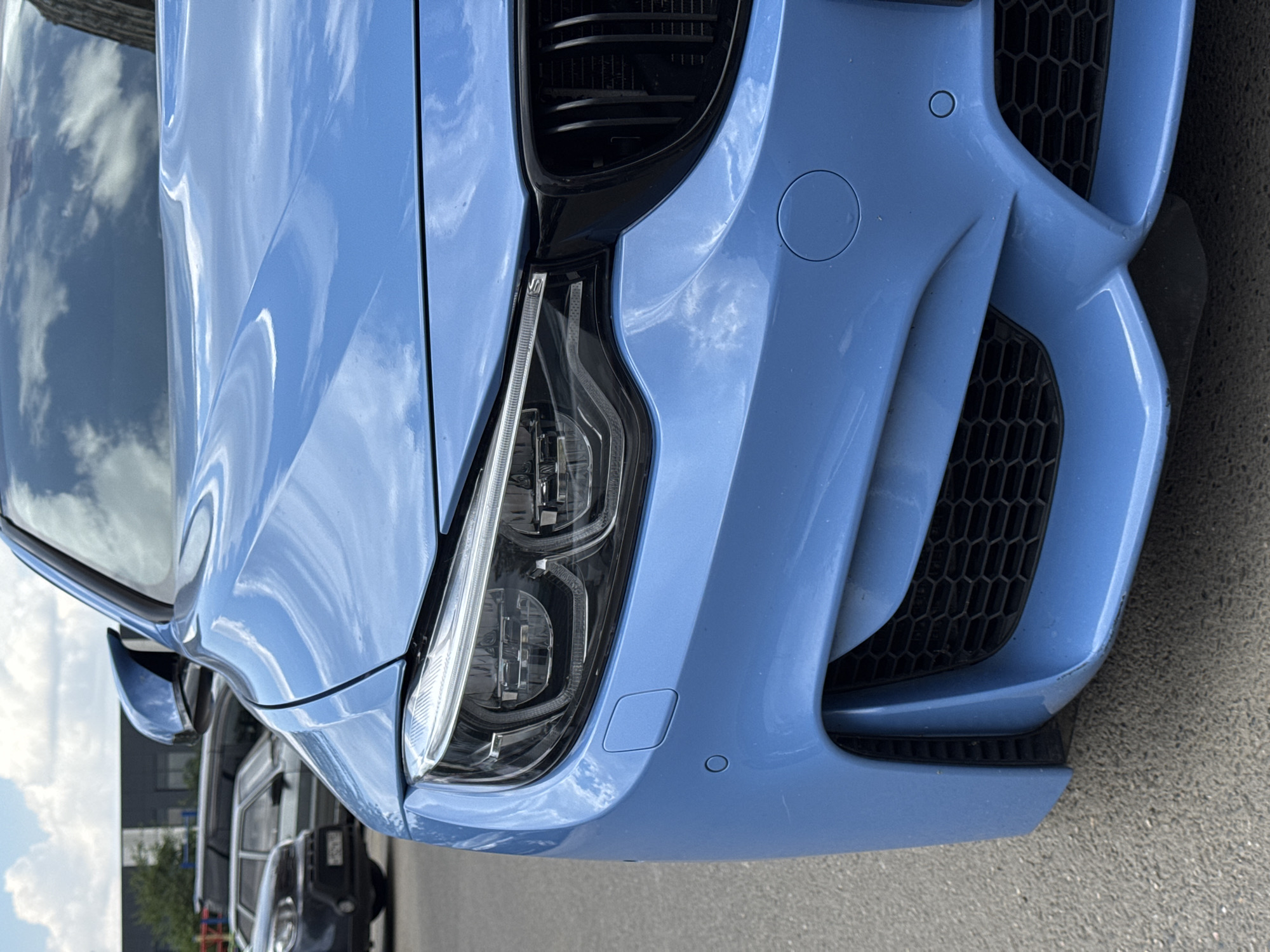 The image shows a close-up, low-angle view of the front left side of a bright blue BMW M4. The car is parked on asphalt, likely in a parking lot or on a street. In the background, partially visible, are other cars, including a dark gray SUV and a light-colored sedan, and a building with a dark facade. The sky above is blue with scattered white clouds. The car's headlight, grille, and part of the hood and bumper are in sharp focus. There are no people visible in the image. The scene appears to be an outdoor urban environment.