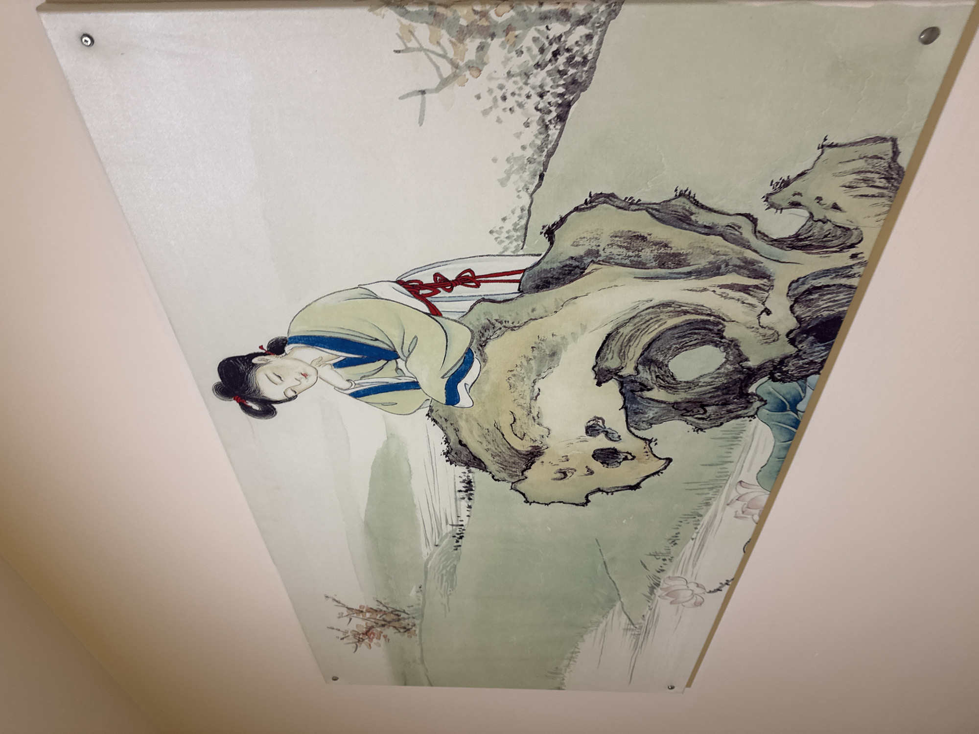 The image shows a painting on a wall, depicting a traditional Chinese scene. In the foreground, a woman in historical Chinese attire is leaning on a large, weathered rock. She has her eyes closed and appears to be in a contemplative pose. The rock is intricately detailed with dark ink strokes, suggesting a rough texture and age. Behind the woman and rock, the landscape features rolling hills painted in soft green washes, with subtle details of trees and possibly a path with figures. In the lower left portion of the painting, lotus flowers are depicted. The painting is a wide rectangular format and is mounted on a wall. The wall itself is a pale pinkish-beige color. There are visible screws or fasteners holding the painting to the wall at its corners and along the top edge. The perspective suggests the photograph was taken from a slightly elevated angle, looking down at the painting. The overall style of the painting is reminiscent of classical Chinese ink wash art.