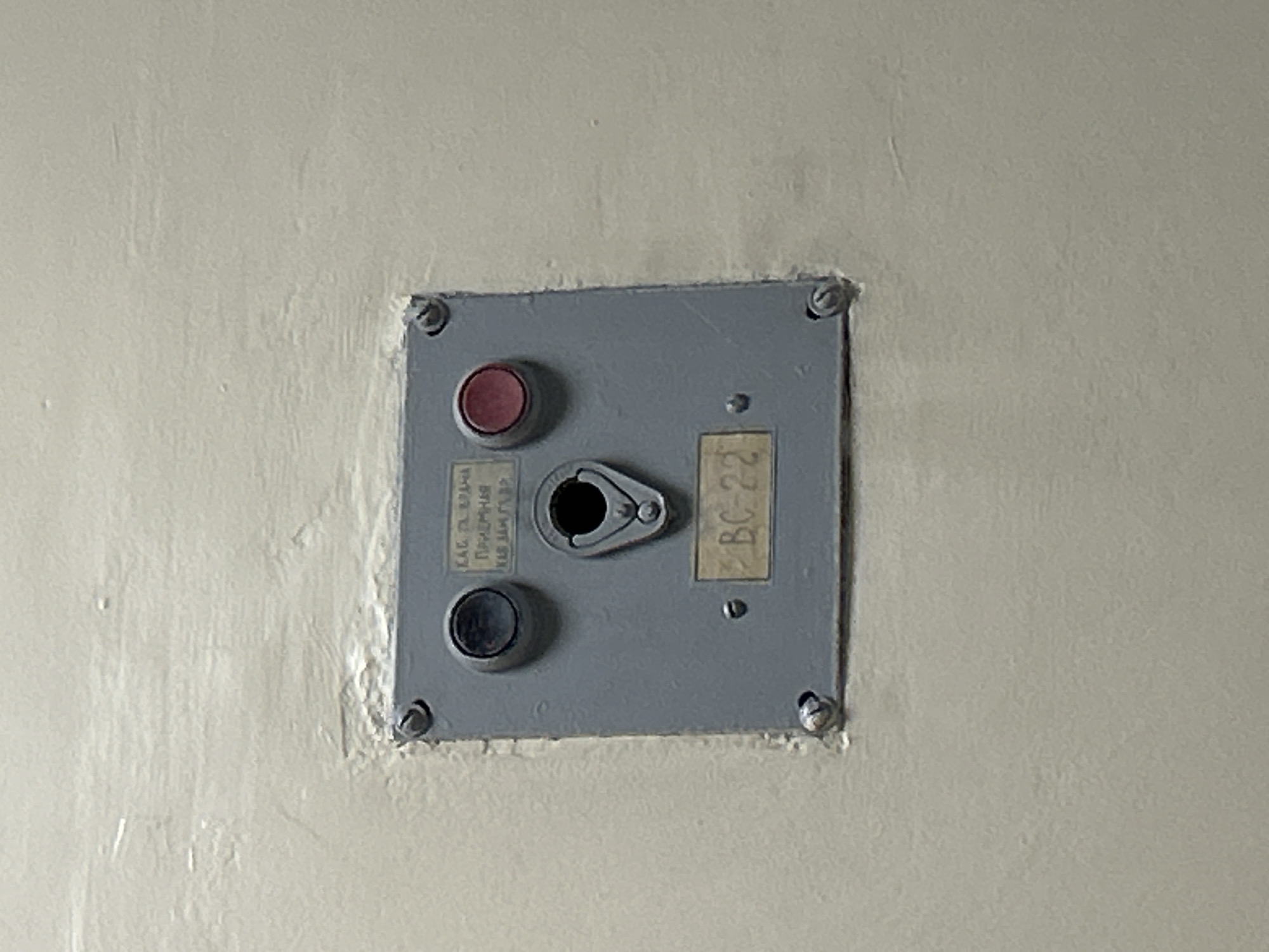 A close-up, eye-level, medium shot captures a gray electrical panel mounted on a textured, off-white wall in Lyubertsy, Russia. The panel, approximately square, is secured by four screws at its corners. In the upper left of the panel is a dark circular button, and to its right, a red circular button. Centered on the panel is a black, hole-like aperture with a metallic surrounding, designed to receive a key or plug. Below this aperture, a rectangular label with black lettering on a beige background reads "BC-22". Above the aperture, a smaller, faded label in Russian appears to have the text "КАБ. ЭЛ. ВРАЧА ПРИЕМНАЯ КАБ. ЗАМ. ГЛ. ВРАЧА". The wall surrounding the panel is uneven and appears to have a rough, stucco-like texture. The lighting suggests an indoor environment, possibly a hallway or utility room. No individuals are present in the frame.