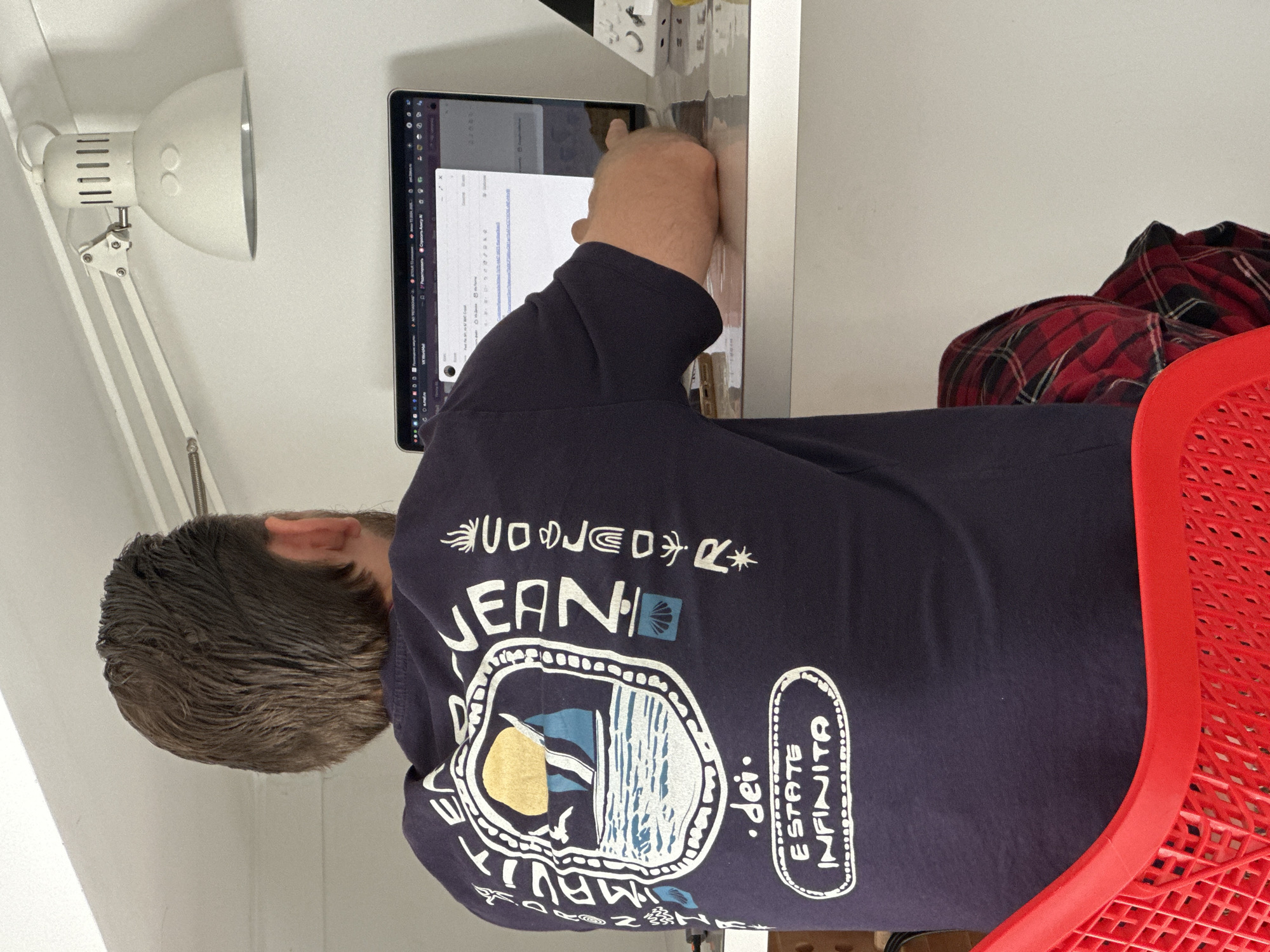 A person is seated at a white desk, viewed from behind, interacting with a laptop or tablet. The individual wears a dark blue t-shirt with several white graphics and text. Visible text on the shirt includes "UOJED", "JEAN", "dei.", and "ESTATE INFINITA" (Italian for "Infinite Summer"), along with a graphic depicting a beach or ocean scene with a flag and stylized waves. The person's hair is dark brown and appears slightly wet or textured.
The laptop screen displays what looks like an email client or document editor with various tabs and elements. Russian text fragments are visible, such as "Яндекс.Почта" (Yandex.Mail), "Отправить" (Send), "Редактировать" (Edit), "Срочно" (Urgent), and "VK Видео" (VK Video), alongside some English terms like "Gmail". The person's right hand is visible, positioned over the device's keyboard or trackpad.
To the left, a white articulated desk lamp with a conical shade extends from the desk. Behind the person, a bright red, textured plastic chair is visible, part of it covered by a folded red and black plaid shirt or blanket. The setting appears to be an indoor room with plain white walls, suggesting a home office or study area during the daytime. The overall scene depicts a person engaged in computer work. The provided location context is Fethiye, Turkey.