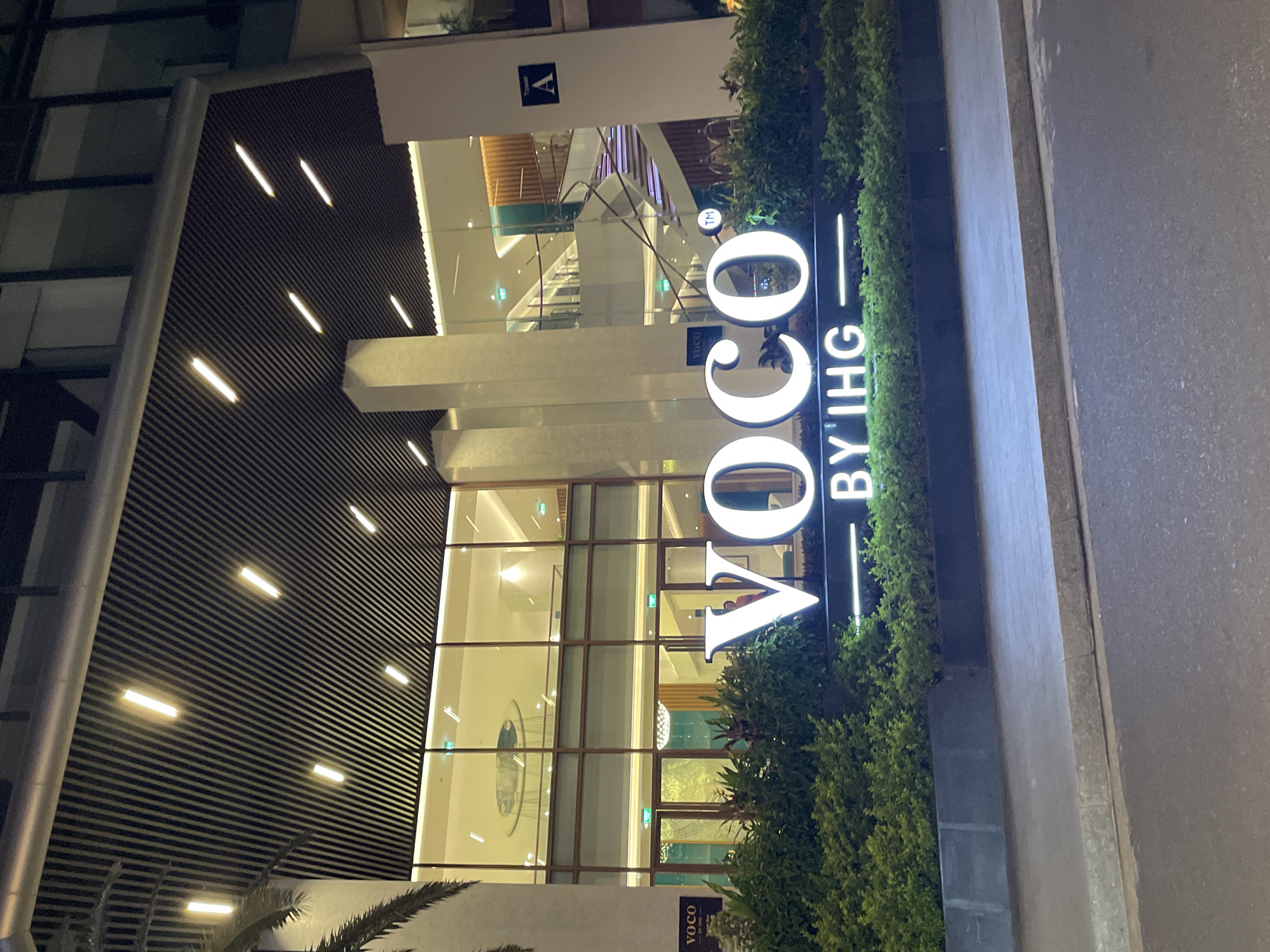 This media file captures the illuminated exterior of a modern hotel building, identified as "VOCO BY IHG," at night in Bắc Nha Trang, Vietnam.
The image shows a multi-story structure with contemporary architecture. To the left, a dark-toned facade features horizontal slat-like patterns and is punctuated by several diagonal linear white lights. The central and right portions of the building are dominated by extensive glass panels, revealing a brightly lit, modern interior. Through these windows, multiple levels, staircases, railings, and what appears to be a large, circular light fixture or art piece can be seen. A small square sign bearing the letter 'A' is visible inside, near the top of the interior.
A large, prominent, vertically oriented sign stands to the right of the building, brightly illuminated with white letters spelling "VOCO" above "BY IHG," separated by a horizontal line. A small "TM" (trademark) symbol is visible next to the 'O' in VOCO. At the bottom right, another smaller sign also displays "VOCO by IHG."
Along the base of the sign and building, a neatly trimmed hedge or row of green bushes adds a touch of nature. In the immediate foreground, a concrete curb or sidewalk runs alongside a dark paved surface, likely a road. A palm frond is partially visible at the bottom left. The overall scene is devoid of people or specific activity, focusing on the static, well-lit architecture against the dark night sky.
