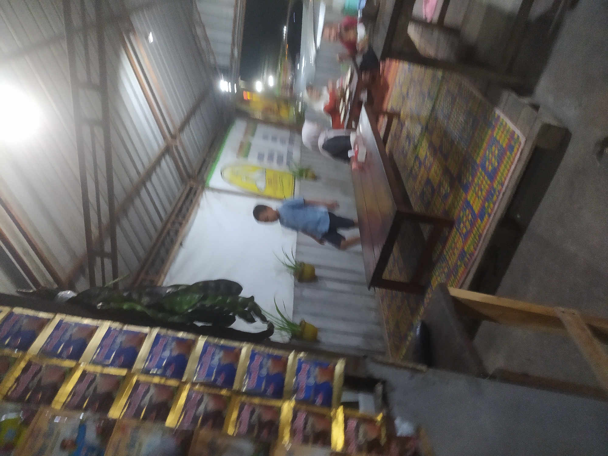 The image captures an indoor scene, likely a warung or small eatery, in Tuntang, Indonesia. The setting appears to be a covered outdoor area with a corrugated metal roof supported by wooden beams. In the foreground on the left, a vertical display rack holds numerous packaged goods, possibly snacks or instant beverages, with prominent blue and gold branding. To the right of the rack, a large, dark green vine hangs down, its leaves partially obscuring the background.
In the mid-ground, a young boy wearing a blue t-shirt and dark shorts stands looking towards the right. Behind him, a white wall serves as a backdrop, with two potted plants hanging at eye level. Further back, another wall features a faded yellow graphic with a stylized human figure. A dark wooden table sits in the center of the frame, placed on a colorful mat with geometric patterns.
To the right of the central table, two women are seated at a smaller table. One woman, wearing a white hijab and a gray top, is looking down at her hands. The other woman, in a red top, is also seated. The lighting is dim, with a bright overhead light creating a glare in the upper portion of the image. The overall atmosphere suggests a casual, communal space, potentially a local gathering spot. The presence of packaged goods and simple seating arrangements points to a typical Indonesian warung.