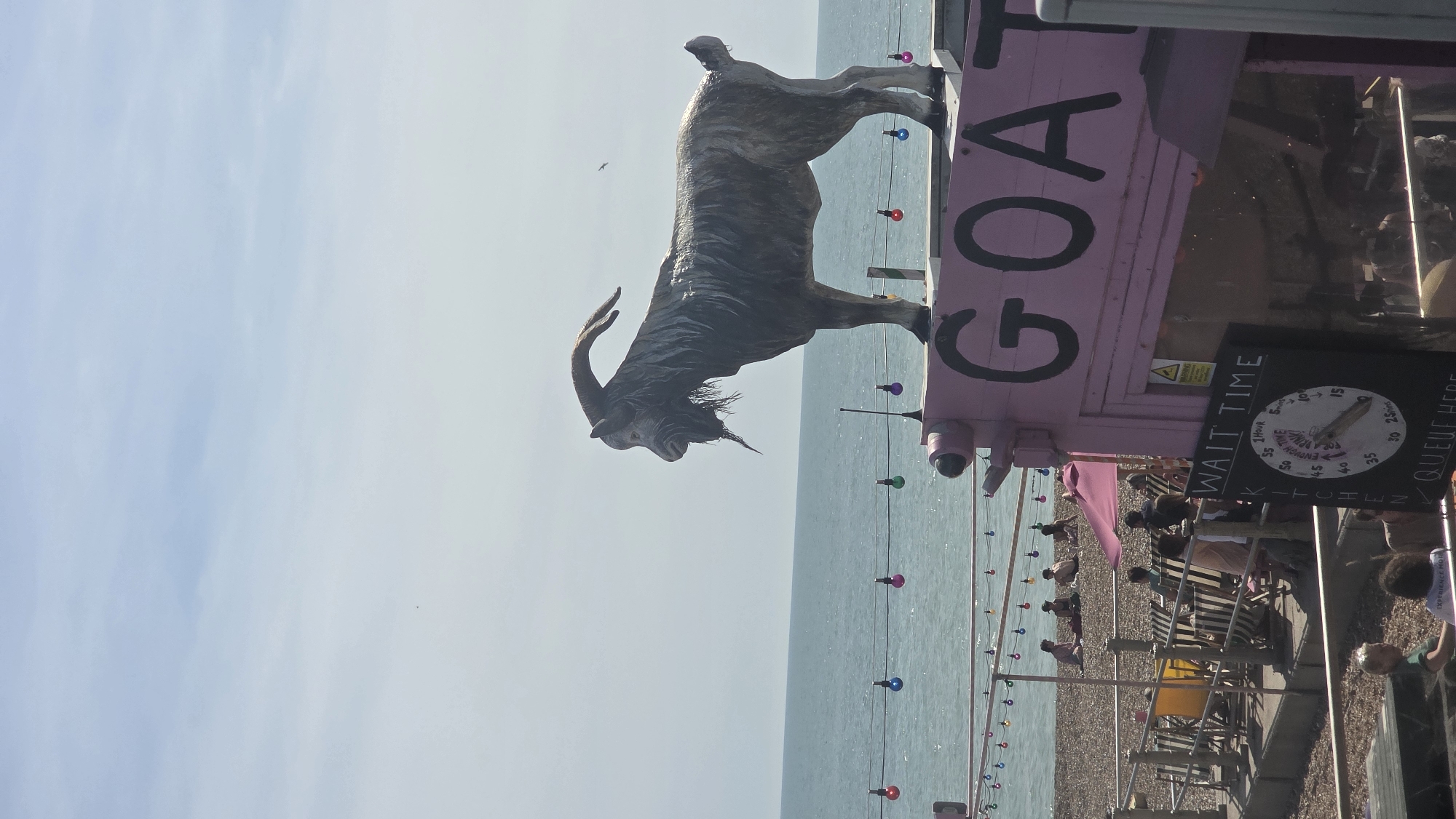An outdoor scene is captured on a sunny day at the beach in St. Leonards, United Kingdom. A large, dark sculpture of a goat stands prominently on top of a pink structure, which appears to be a stall or building with the word "GOAT" painted in large black letters on its side. The goat sculpture is detailed, showing its horns, beard, and textured fur.
Below the goat sculpture and the pink structure, a beach is visible, populated with people lounging and sitting. Deck chairs and umbrellas suggest a recreational area. The sea stretches out behind the beach, with a clear horizon. A string of colorful lights is strung across the foreground, adding a festive atmosphere.
In the lower right portion of the image, a sign displays "WAIT TIME" with a circular dial indicating various waiting periods, from 25 minutes to 1 hour, and a note that reads "ENOUGH TIME FOR A DRINK". This suggests the pink structure is likely a food or drink establishment. People are also visible in the lower part of the image, some seated, suggesting they are patrons. The overall impression is a lively beachside scene on a bright day.