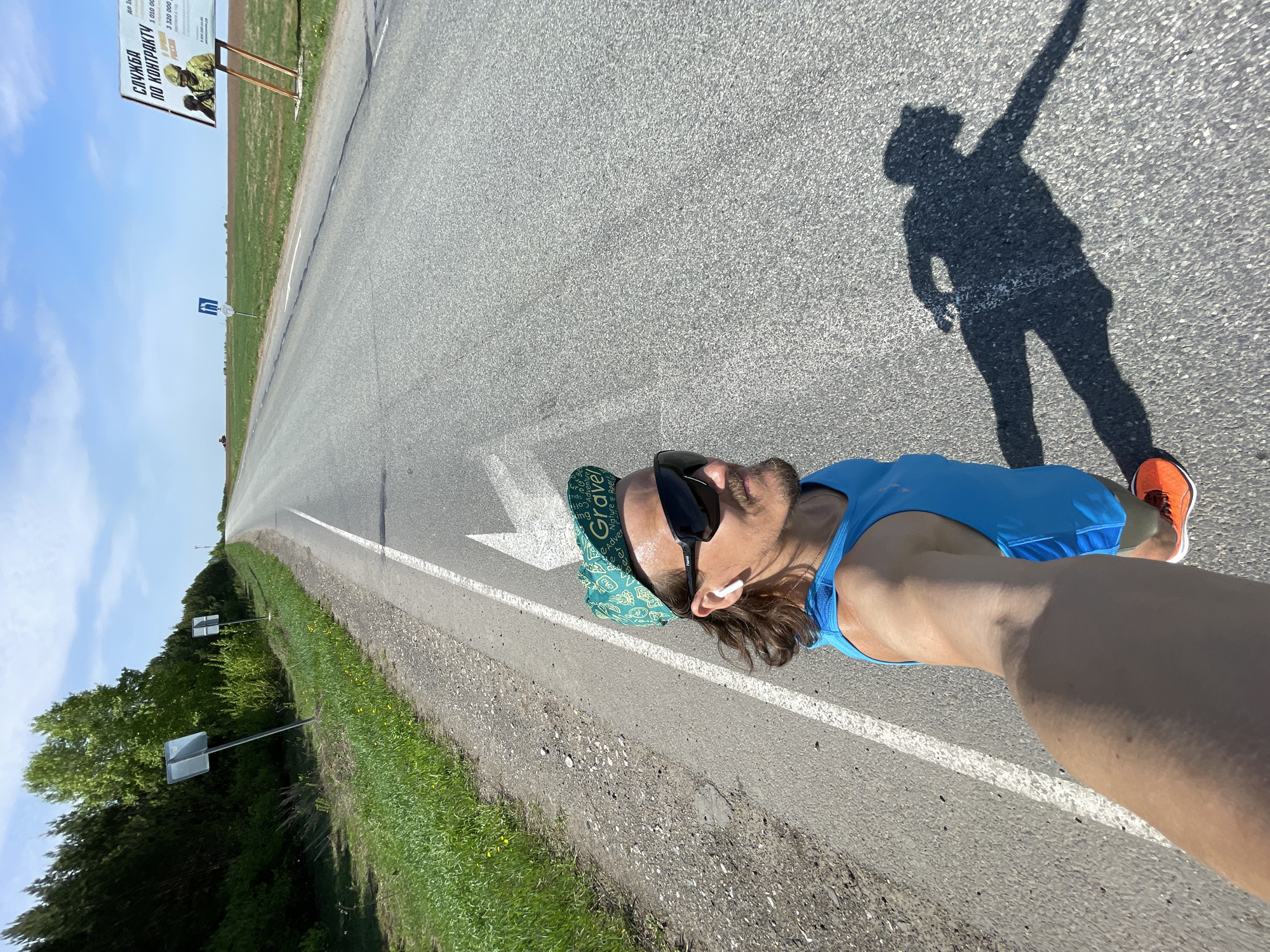 The image shows a man taking a selfie on a road, likely while running. He is wearing a blue tank top, shorts, sunglasses, a bandana, and orange running shoes. A shadow of the man is cast on the road. The road has a white line dividing it. In the background, there is a billboard with Cyrillic writing and vegetation. Based on the text on the billboard, "СЛУЖБА ПО КОНТРАКТУ," and the location information provided, it can be inferred that the image was taken in or near the city of Перевозное, Russia. The billboard seems to advertise military service.
