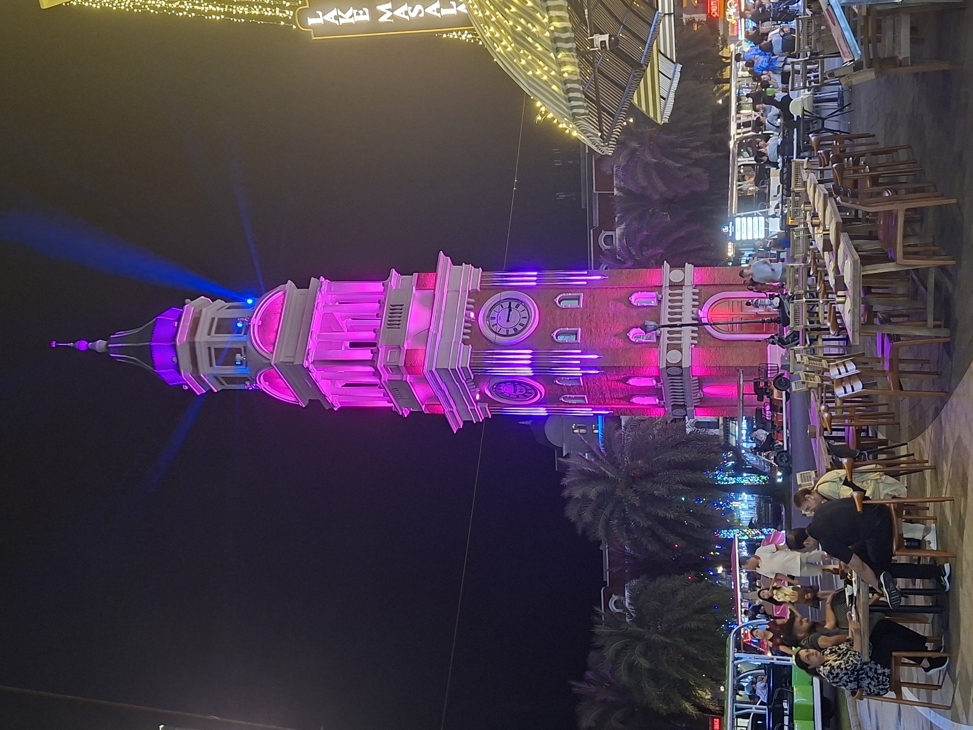 The image depicts a nighttime scene at a bustling outdoor dining area in the city of Phú Quốc, Vietnam. A prominent clock tower, illuminated with vibrant pink and purple lights, stands tall in the background, casting a colorful glow. Palm trees are visible surrounding the tower. In the foreground, several people are seated at tables, enjoying meals and drinks at an outdoor restaurant. The tables are set with plates and utensils. A green tourist shuttle bus is stopped nearby, with people boarding or disembarking. Other people are seen walking around, and more tables and chairs suggest a lively atmosphere. The overall scene captures the ambiance of a popular entertainment or dining district at night.