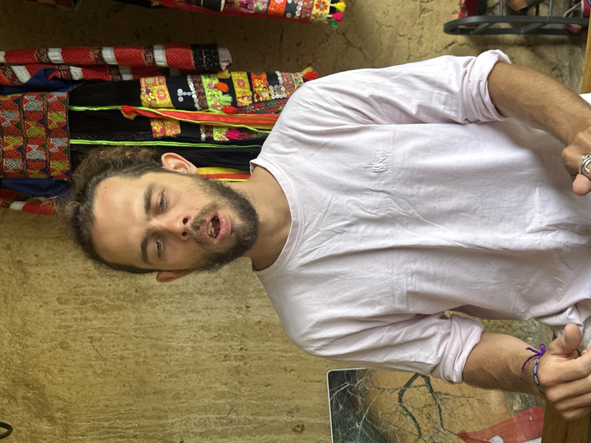A man with dark hair and a beard stands in what appears to be a shop or market stall in Lũng Cú Commune, Vietnam. He is wearing a light pink long-sleeved shirt with the sleeves rolled up. He has a silver ring on his hand and a bracelet on his wrist. His mouth is open, as if he is speaking or gesturing. Behind him are colorful textiles or garments stacked or hanging on display, and there is a beige wall. Below his arm is a picture frame or tablet. The scene suggests he is interacting with someone off-camera, potentially a vendor or customer.